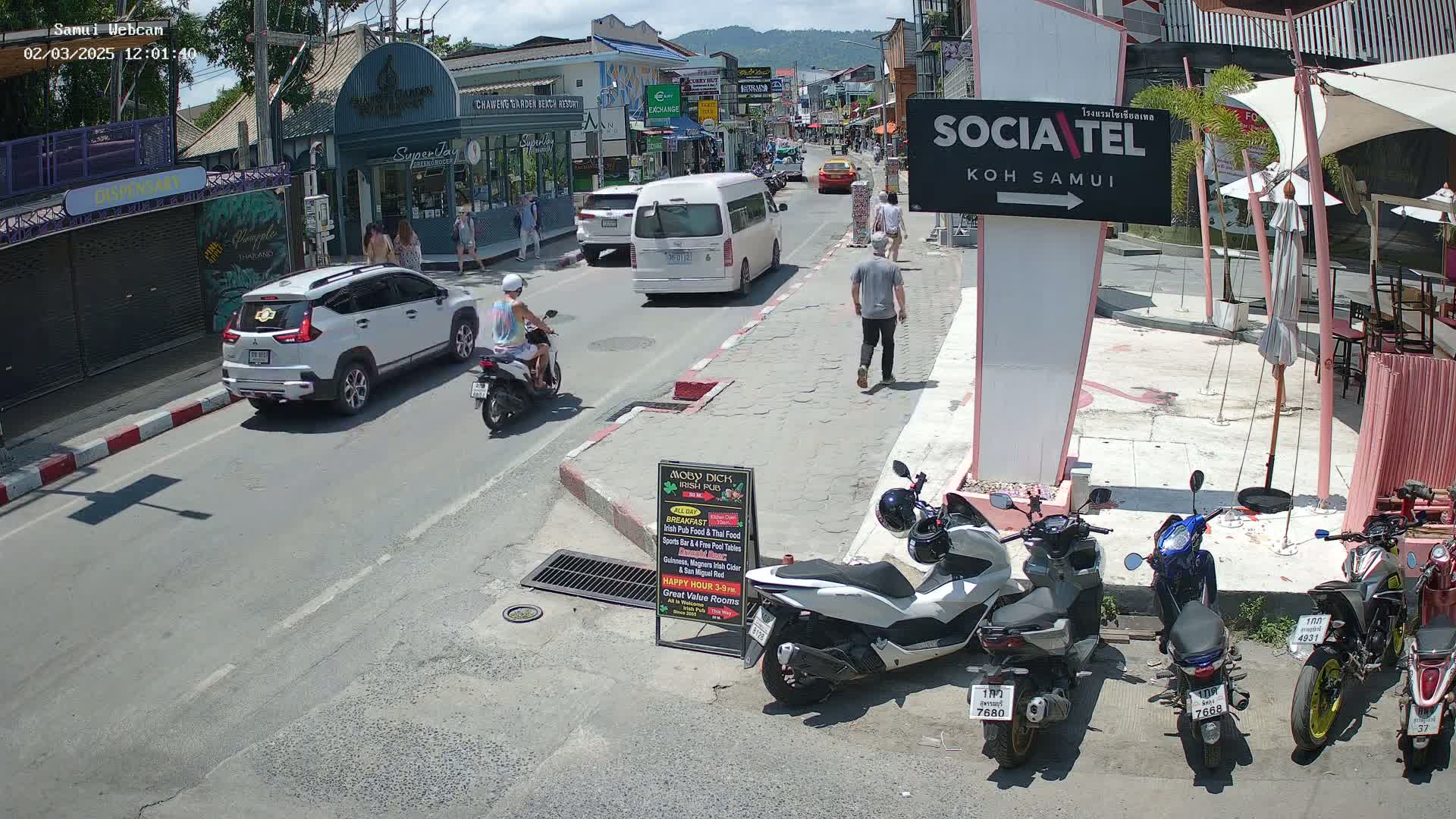 A sunny street scene shows several vehicles, including cars and motorbikes, parked and driving along a road lined with shops and restaurants, with people walking on the sidewalks.