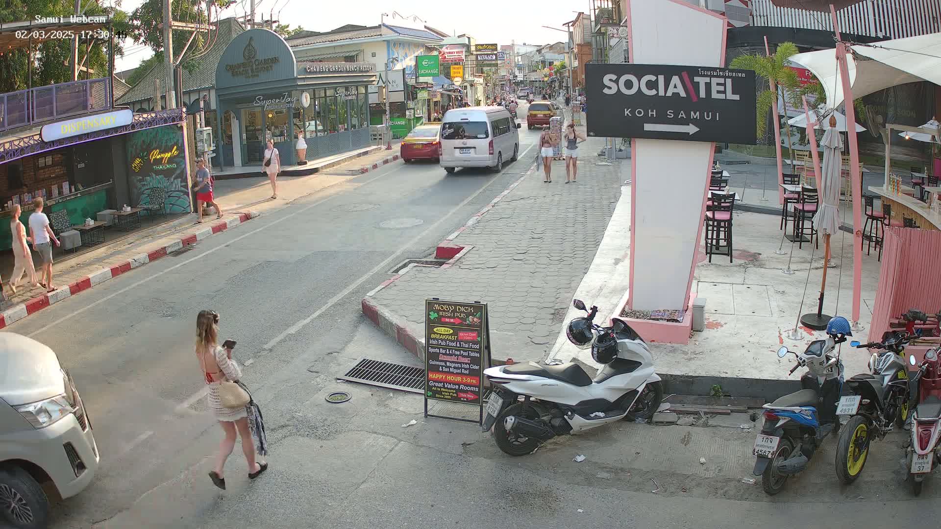 A street scene in a town shows pedestrians walking past shops and restaurants, with several parked scooters and a minivan driving down the road, under sunny evening conditions.