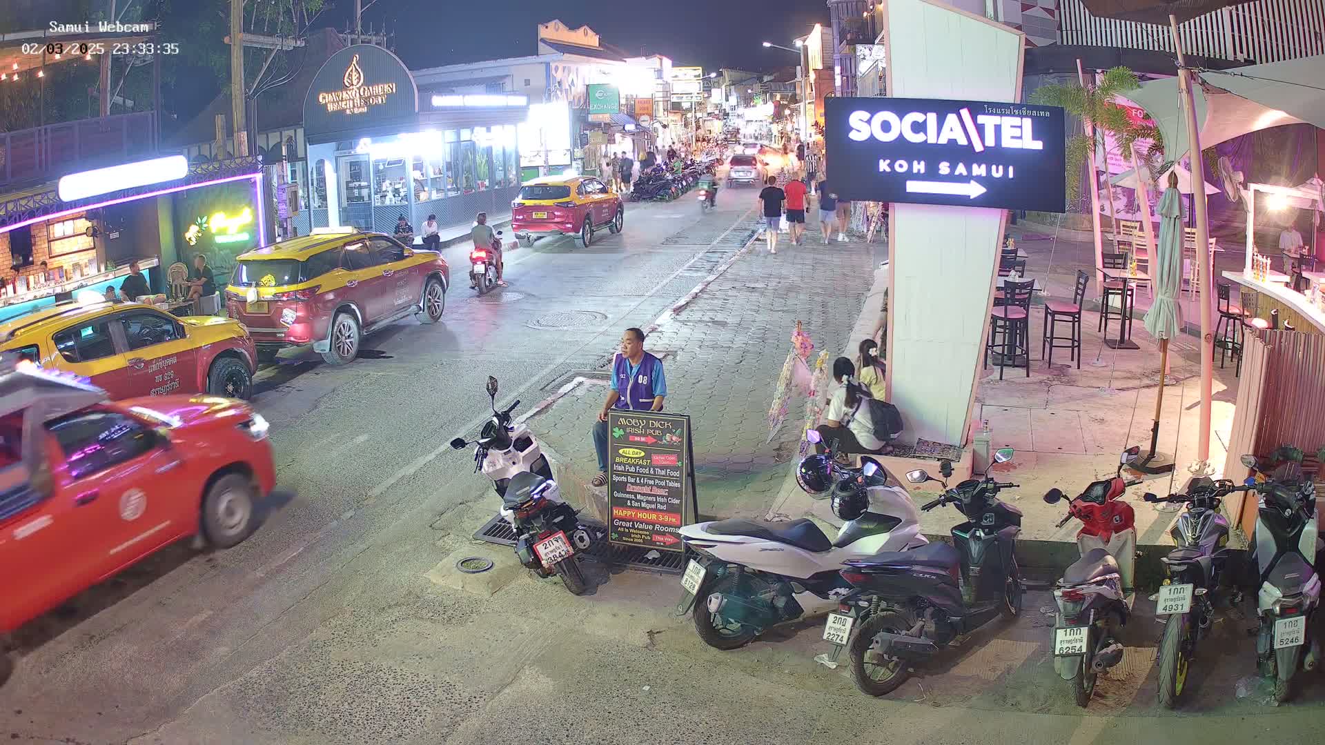 A nighttime street scene shows parked cars and motorbikes, a man holding a menu board, and people walking along a road lined with businesses.