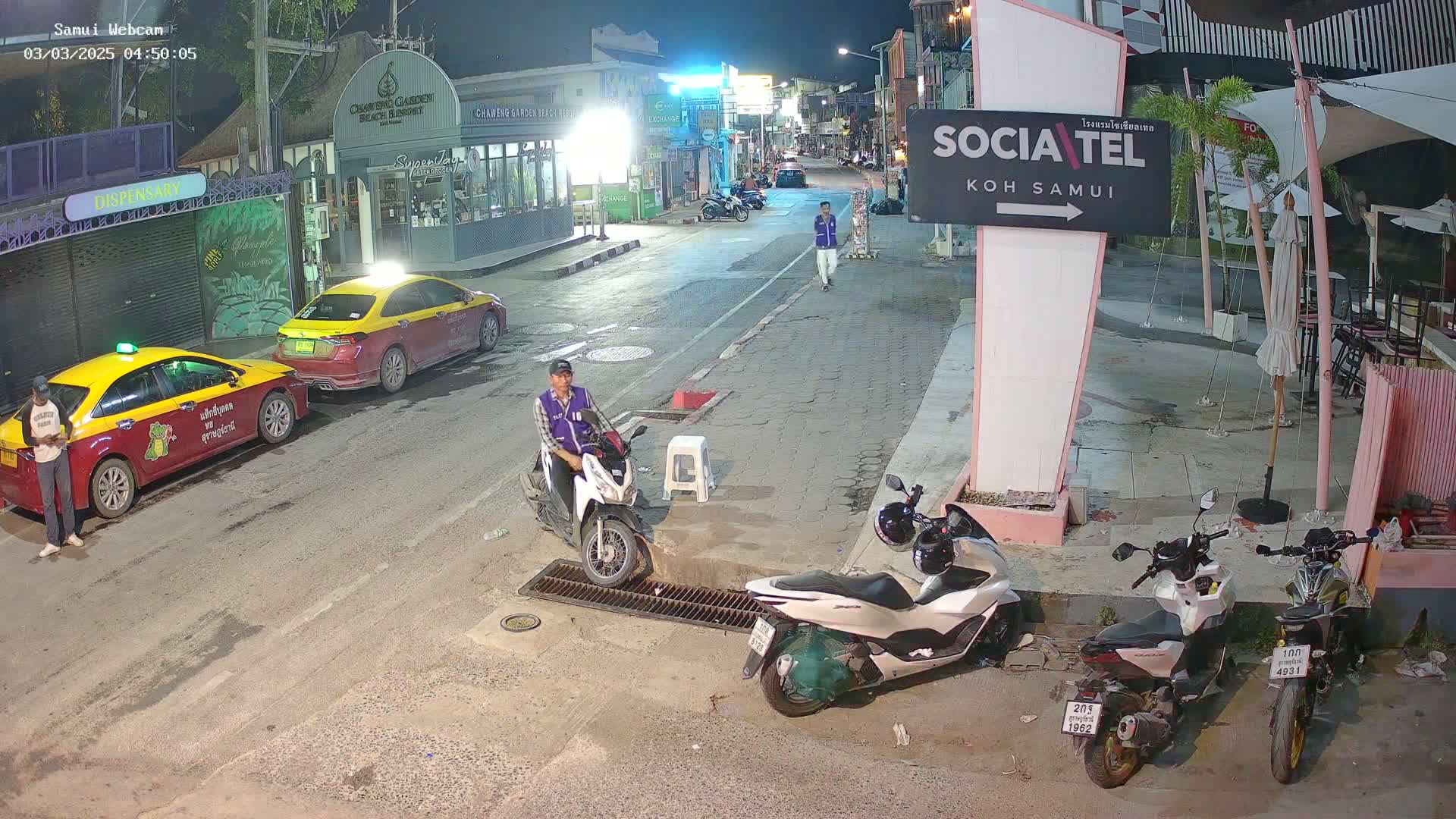 A nighttime street scene shows two taxis parked, a person standing near one, another person sitting on a scooter, and several motorcycles parked nearby.