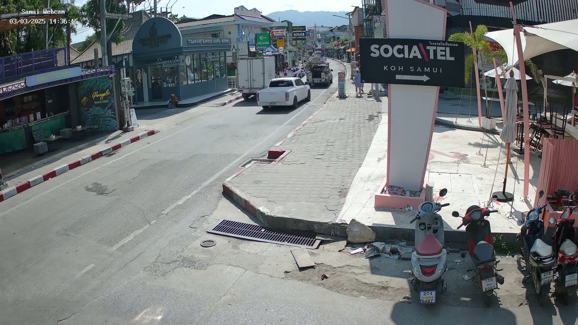 A sunny street scene shows a road with vehicles, including pickup trucks and motorbikes, shops and buildings lining the street, and outdoor seating areas under umbrellas.