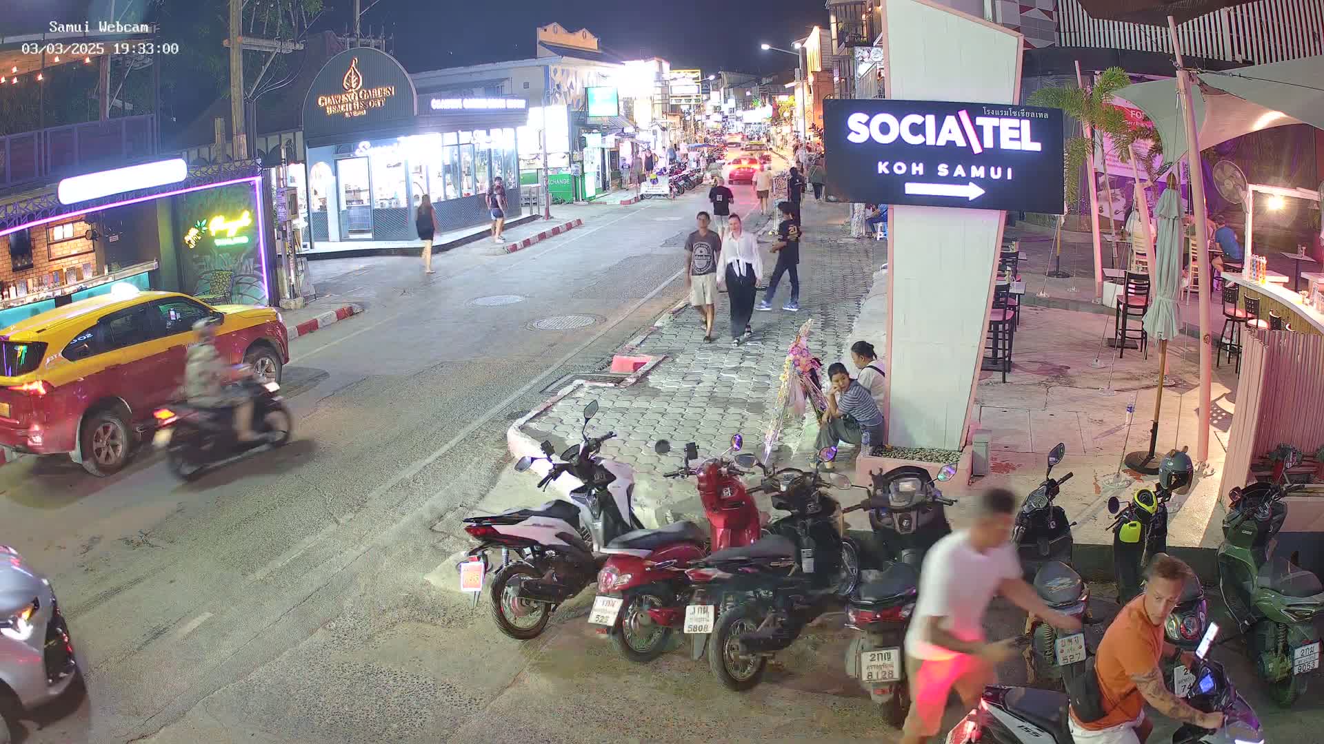 A nighttime street scene shows pedestrians walking past shops and restaurants, with several parked motorbikes and a few cars, under clear skies.