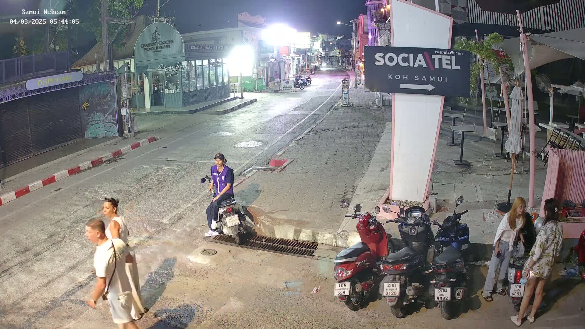 At night, several people stand and walk near parked motorbikes on a street lined with buildings, under dim ambient lighting.