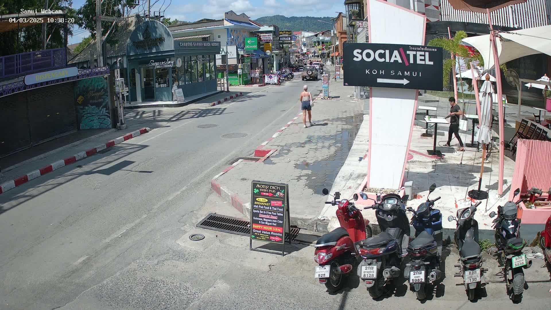 A sunny street scene shows a woman walking down a street lined with shops and restaurants, several motor scooters parked along the side, and a man cleaning outside a cafe.