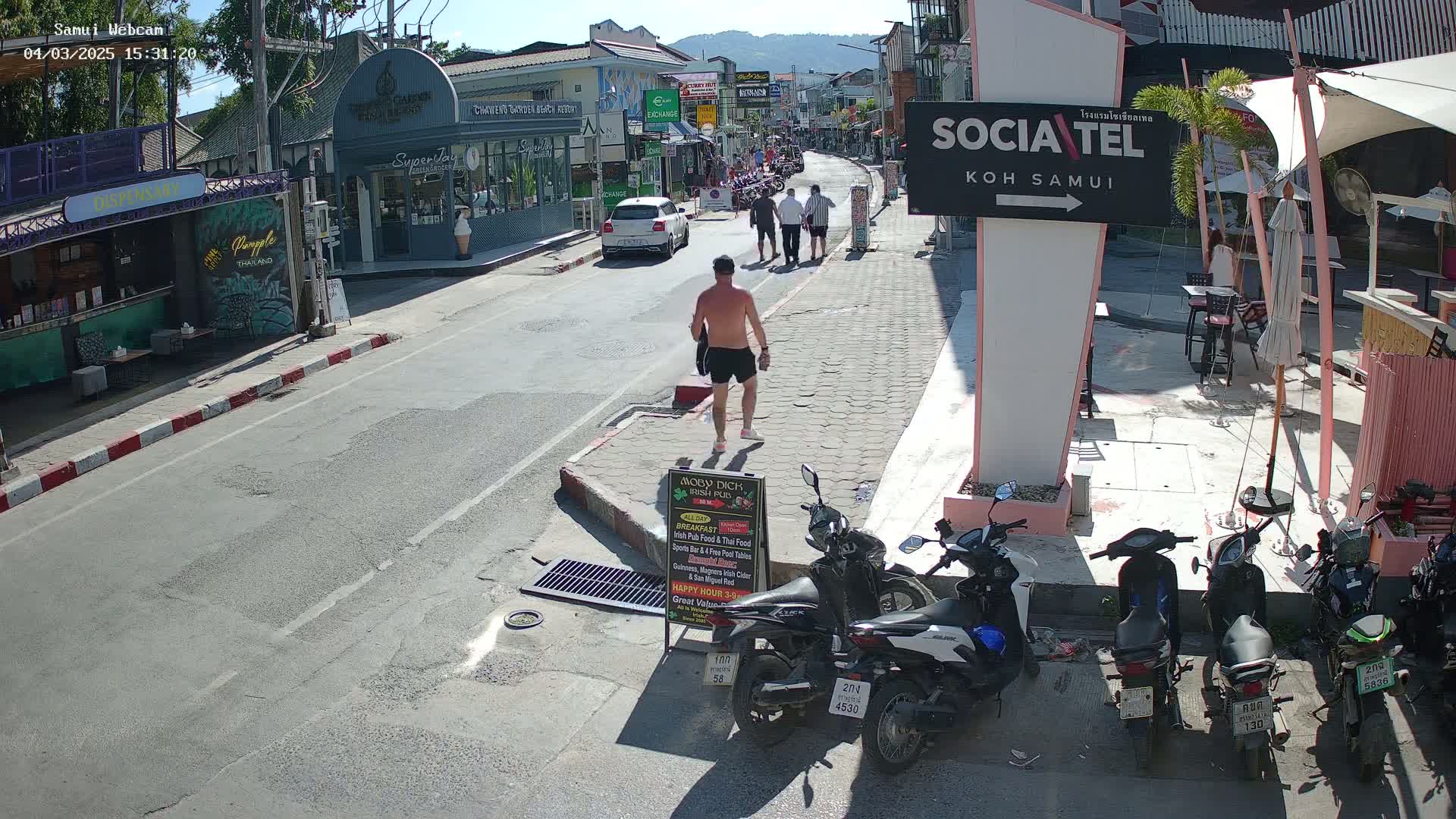A sunny street scene shows a shirtless man walking past shops and parked motorbikes on a partly sunny day.