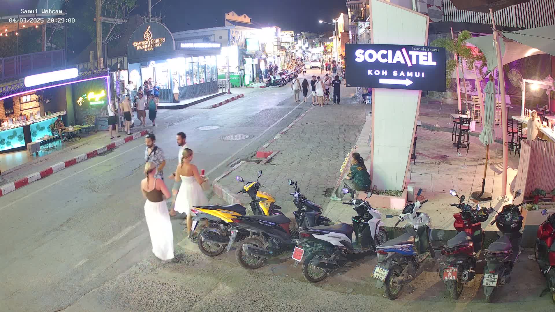 A nighttime street scene shows several people walking and standing near parked motorbikes in front of various businesses, under clear skies.