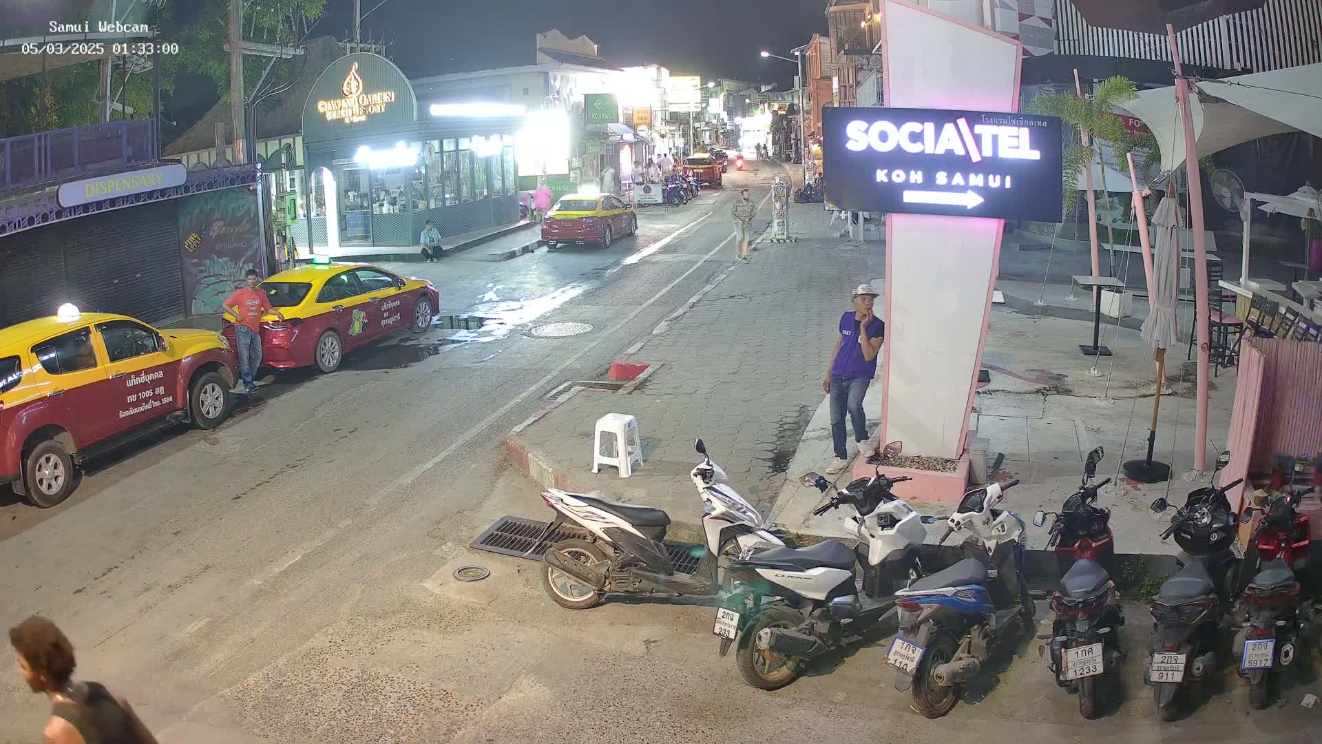 At night, a street scene shows several parked taxis and motorbikes, a few pedestrians, and a man standing near a pink sign in front of a restaurant.