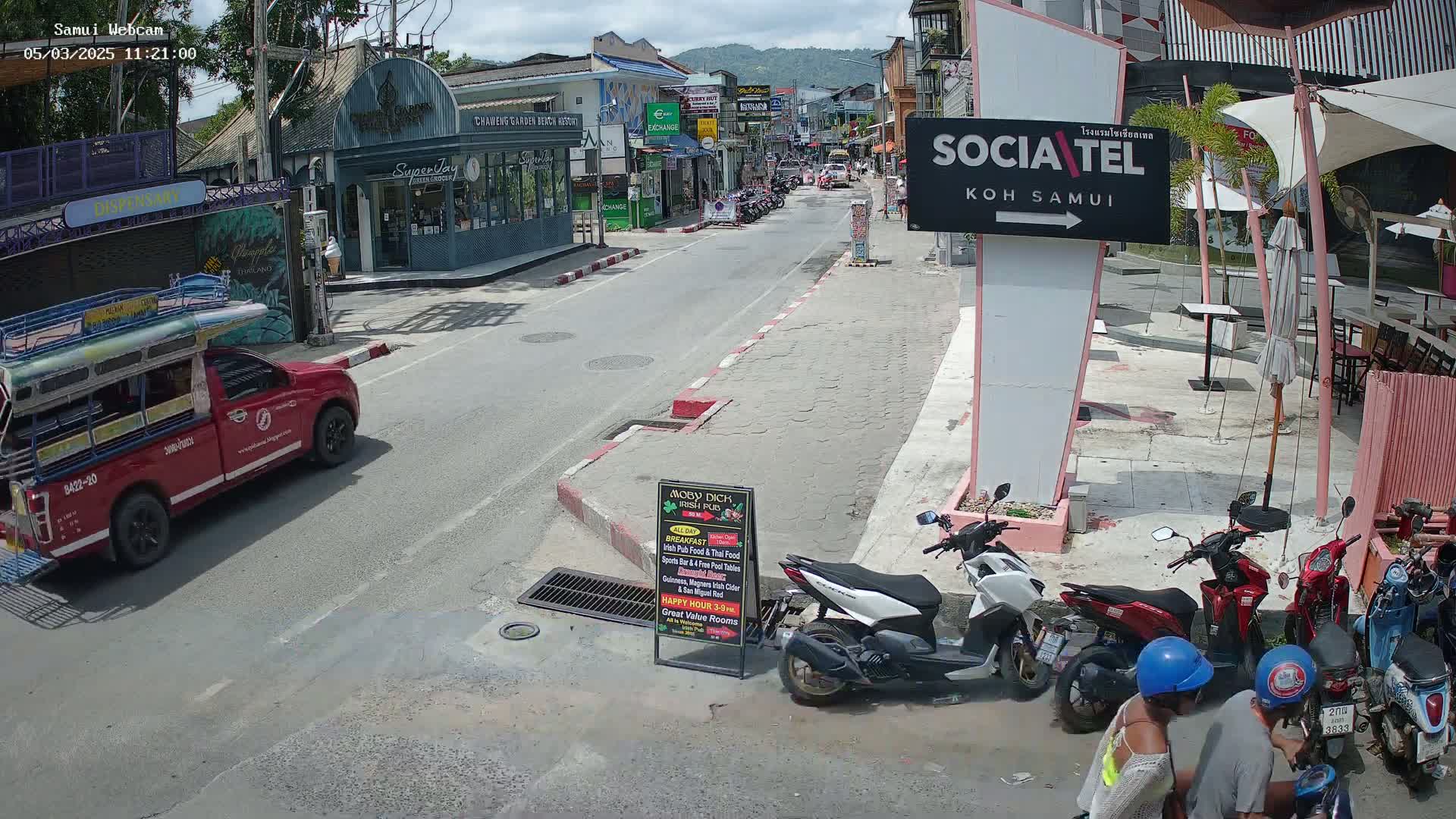 A sunny street scene shows a red pickup truck, several parked motorbikes, and pedestrians near outdoor seating areas, with buildings lining both sides of the road.