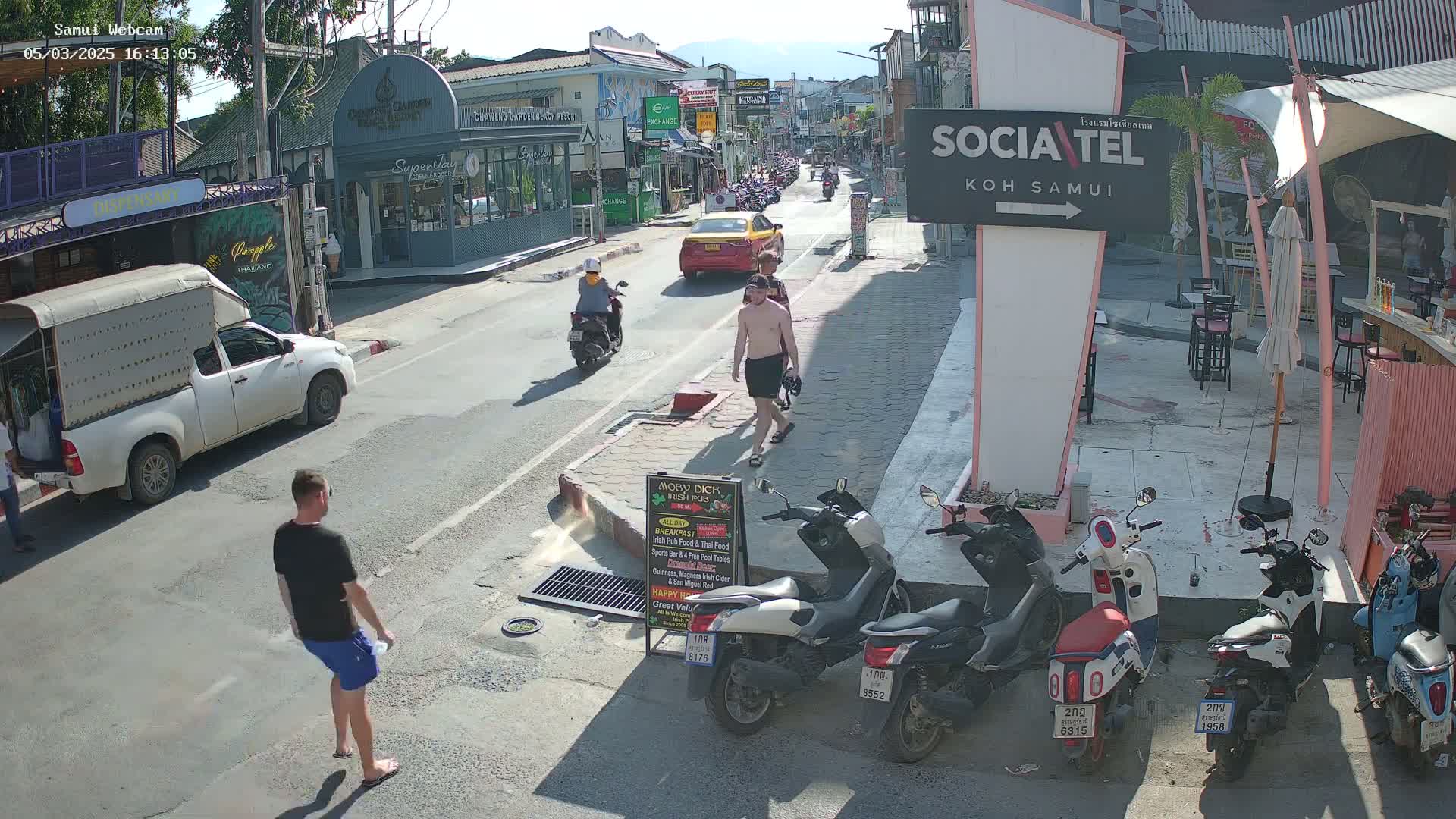 A sunny street scene shows pedestrians and vehicles, including a yellow taxi, motorbikes, a white pickup truck, and several parked scooters, on a street lined with shops and restaurants.