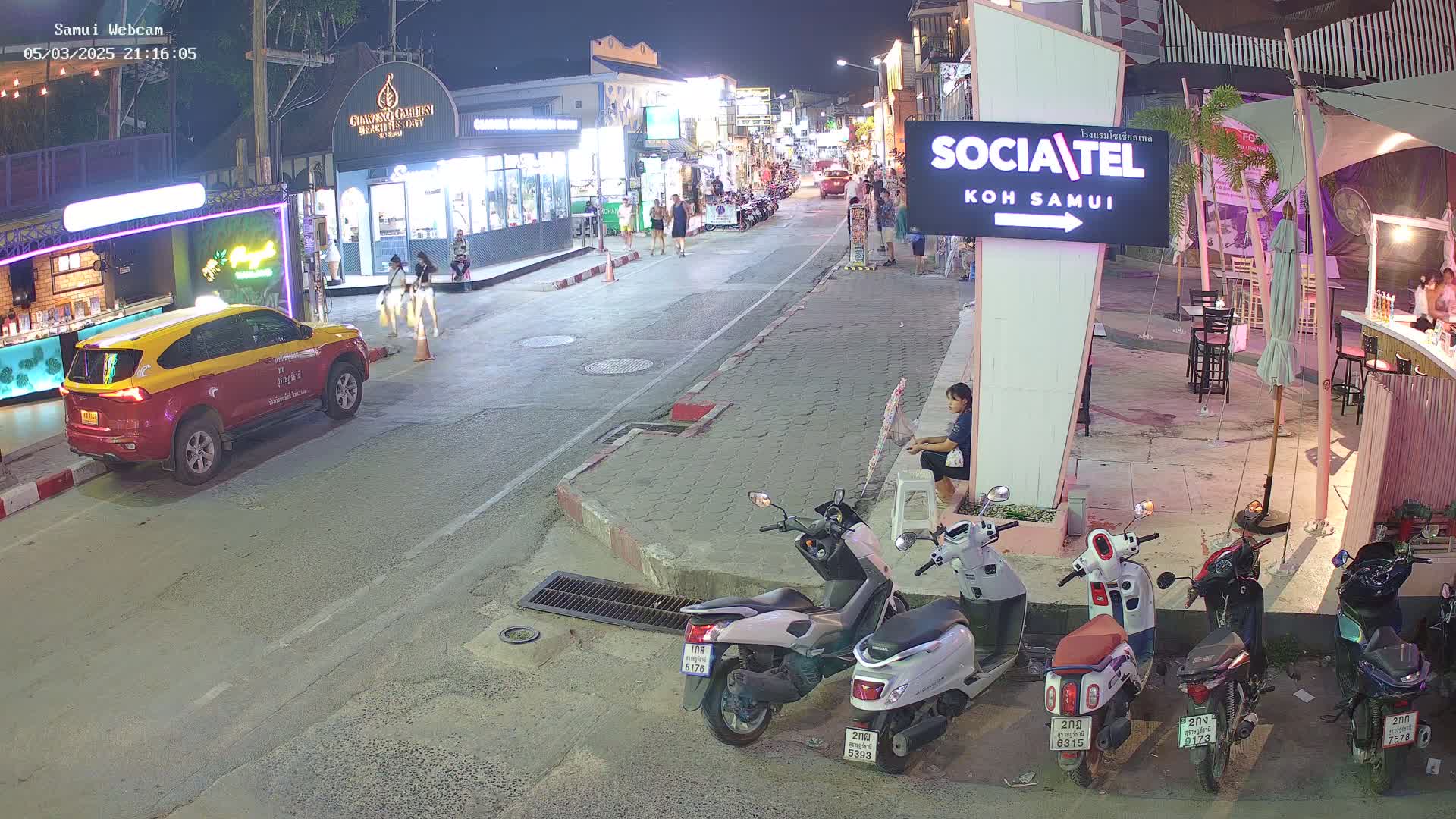A nighttime street scene shows a mostly empty road with parked motor scooters, a taxi, and a few pedestrians, under clear skies.