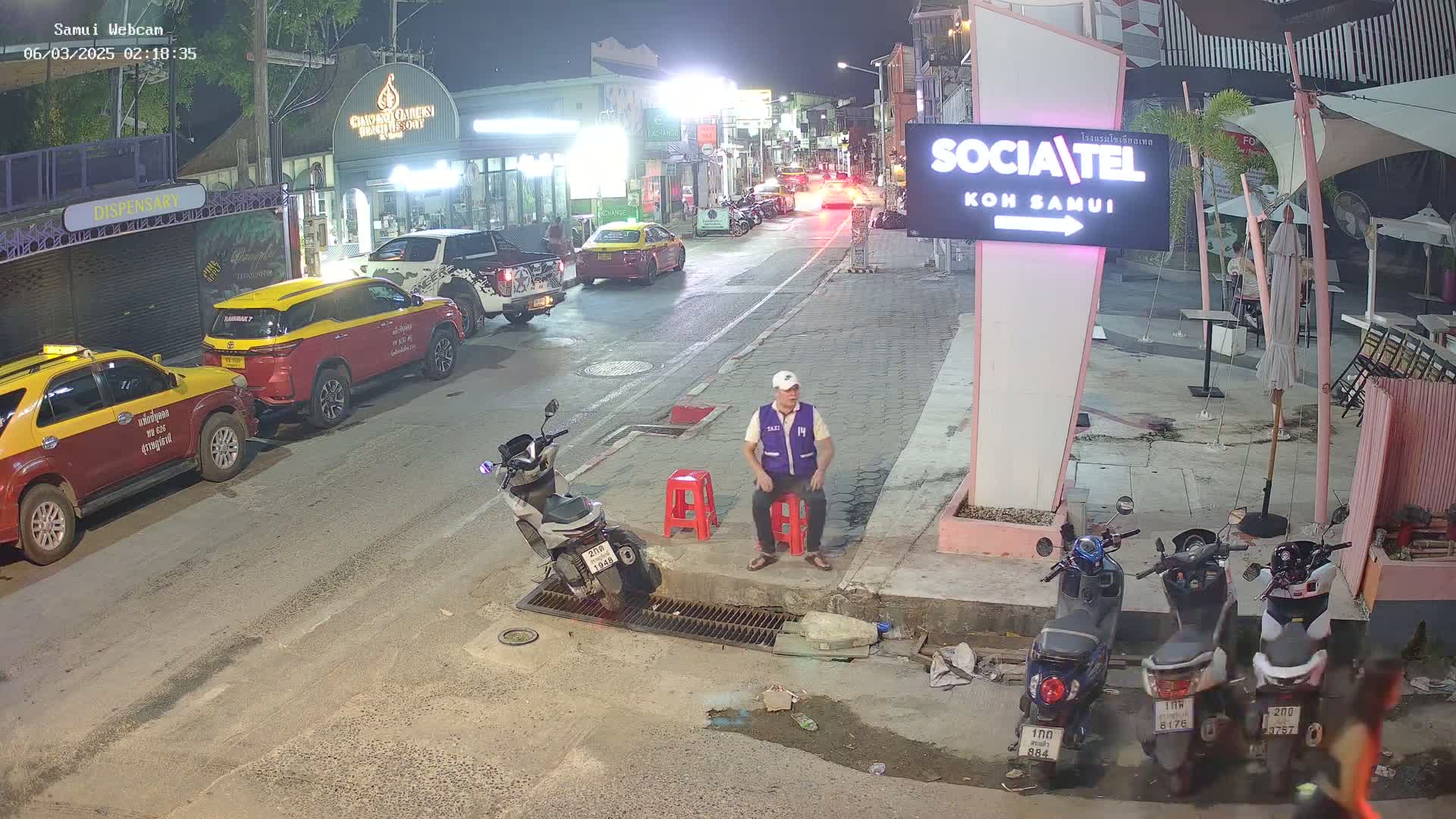 A man in a vest sits on a red stool next to several parked motor scooters on a nighttime street with parked cars and buildings lining the road.