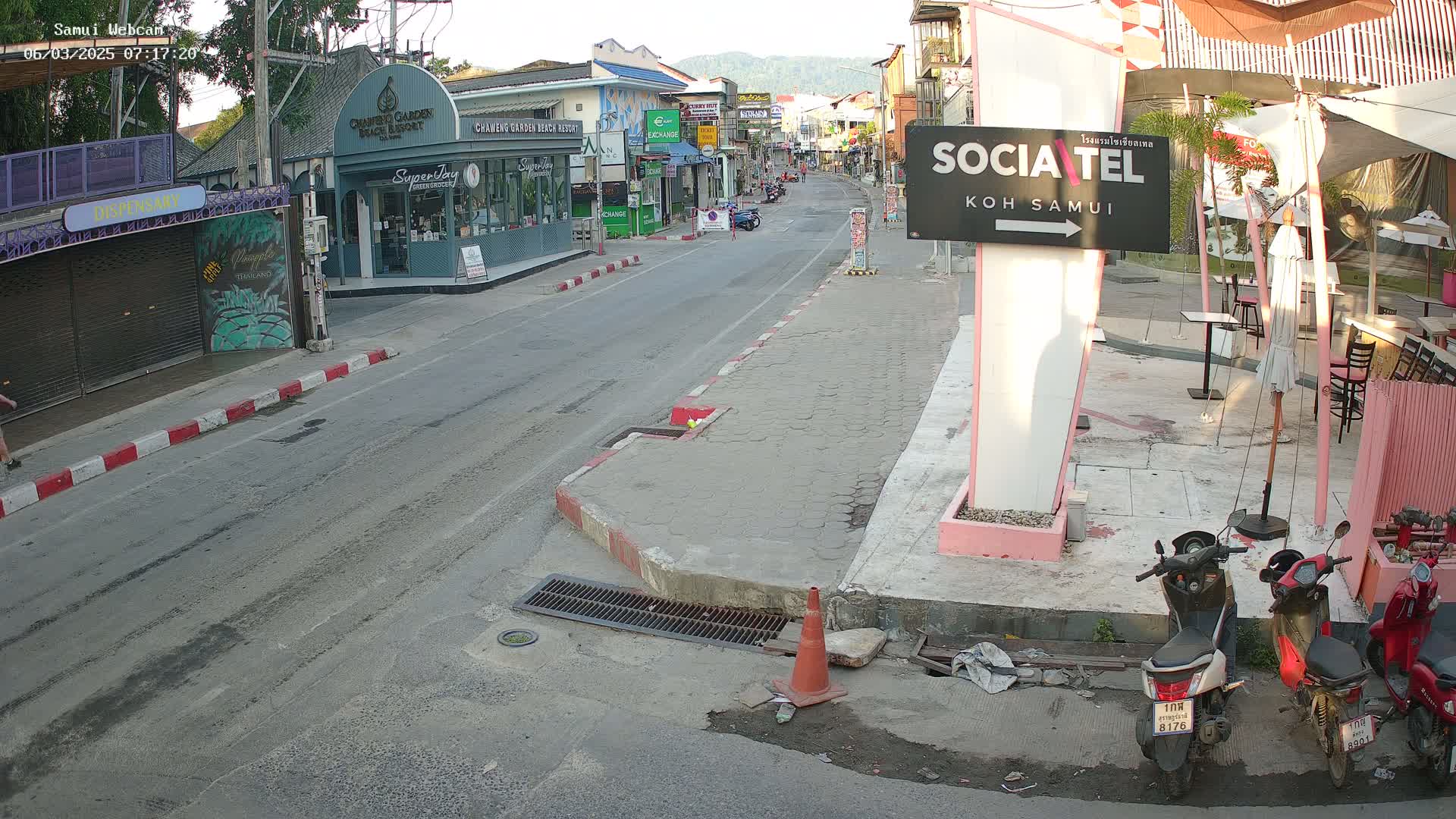 A mostly empty street scene in bright daylight shows shops and restaurants lining the road, with a few parked motor scooters and an orange traffic cone near a pink sign.