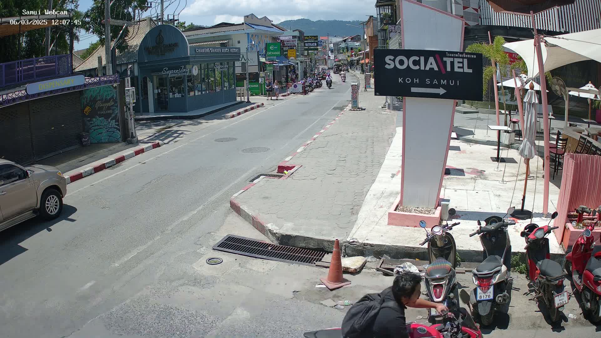 A sunny street scene shows parked motorbikes, a person sitting on a motorbike, and a street with buildings and pedestrians, some of whom are walking along the sidewalk.