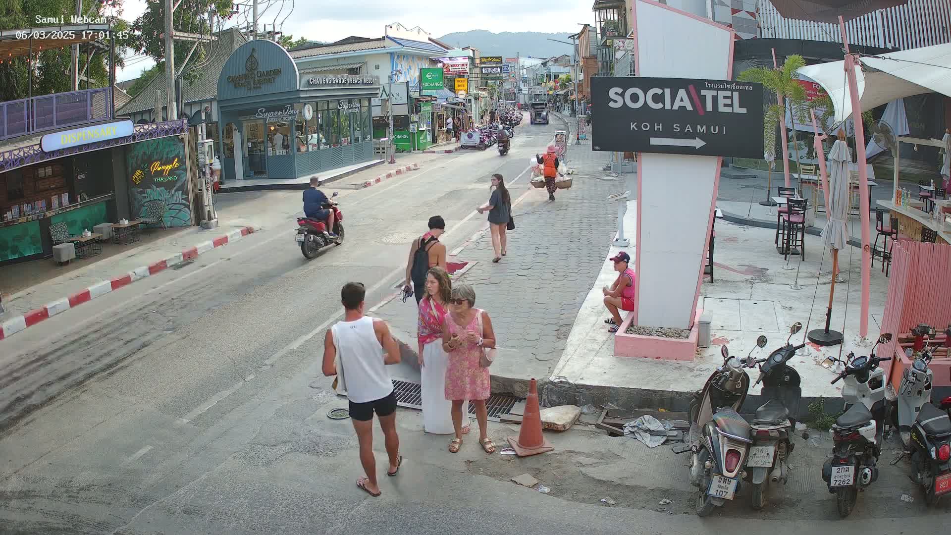 A street scene in the late afternoon shows several people walking and standing near shops and restaurants, with several motorbikes parked along the sidewalk.