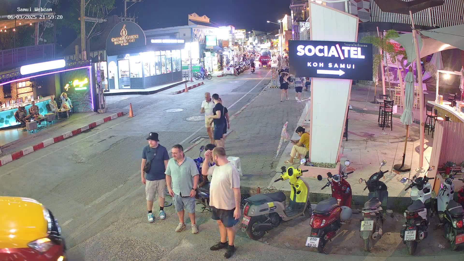 A nighttime street scene shows several people standing and walking near parked motor scooters outside shops and restaurants, under a clear night sky.
