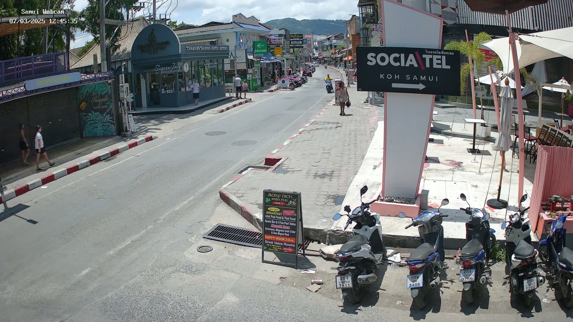 A sunny street scene shows a row of parked motorbikes next to an outdoor cafe, with pedestrians walking along a roadway lined with shops and buildings.
