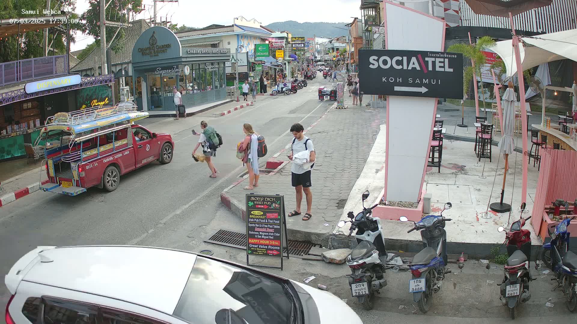 A street scene in the late afternoon shows several pedestrians, a red songthaew, and parked motorbikes, under partly cloudy skies.