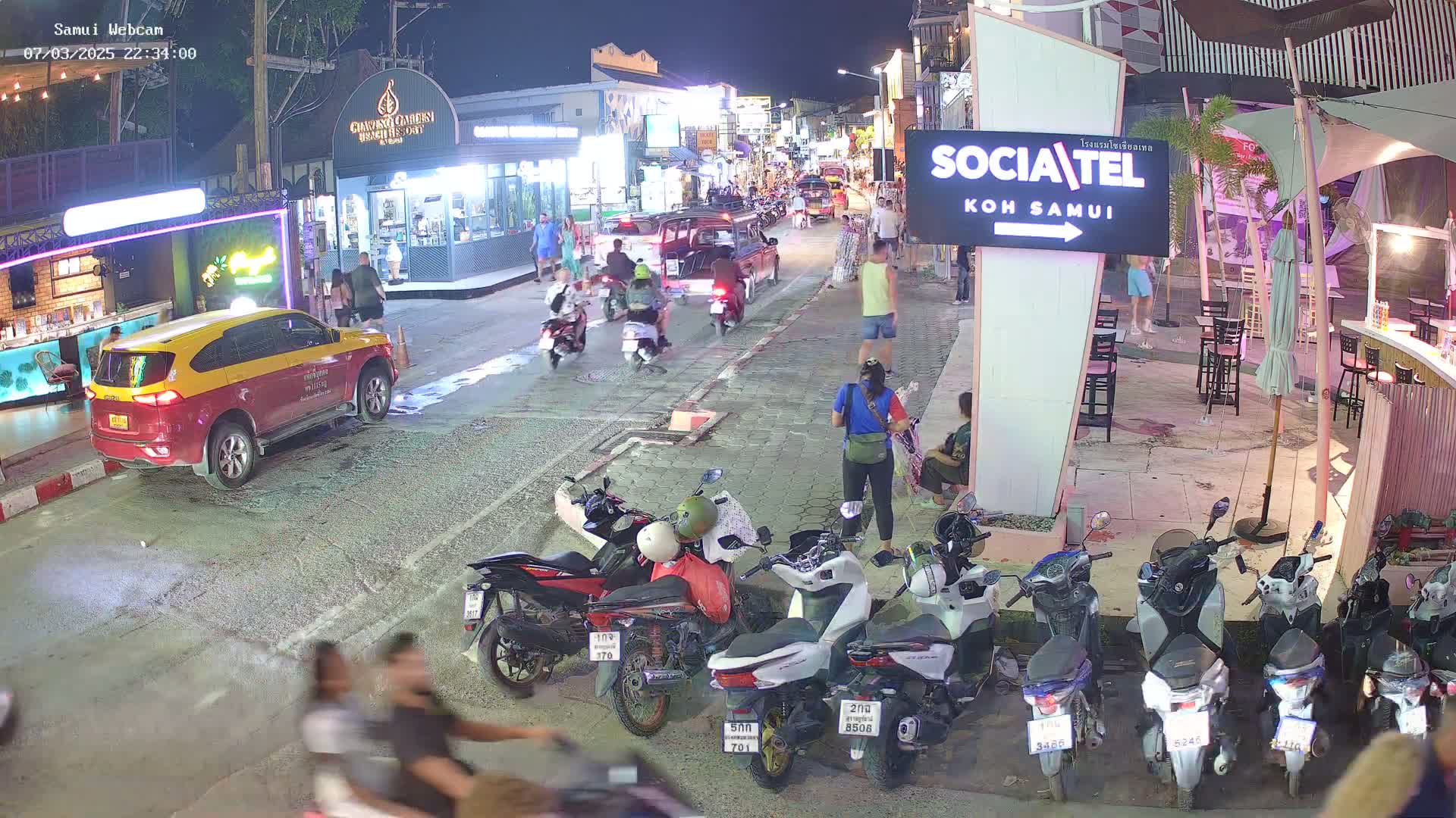 A nighttime street scene shows parked and moving motorbikes and cars, pedestrians, and outdoor seating areas of businesses under clear skies.