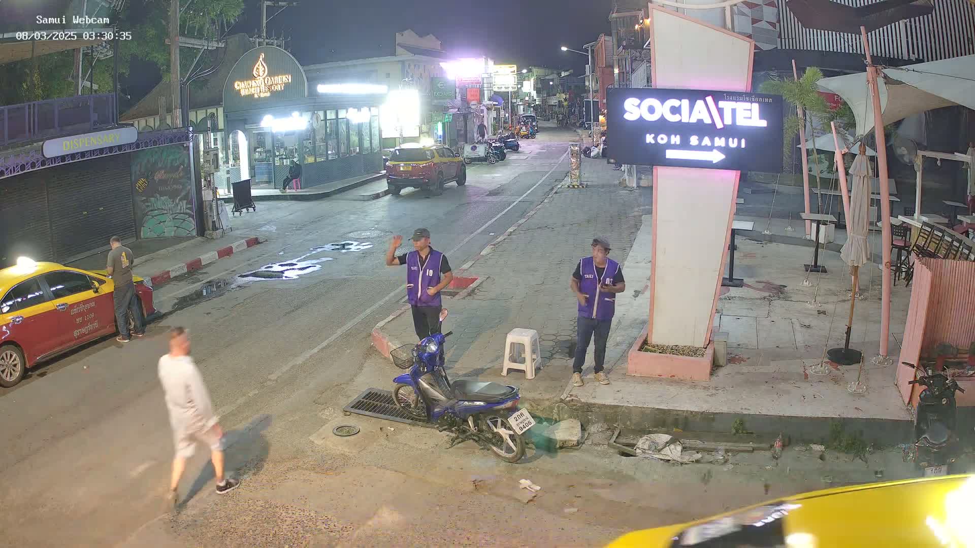A nighttime street scene shows people walking and standing near parked vehicles and a motorcycle, under a clear sky.