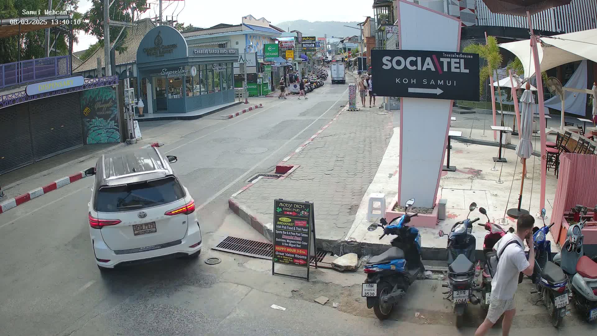 A mostly sunny street scene shows a white SUV parked, several motorbikes parked and a man on a phone, all in front of shops and restaurants.