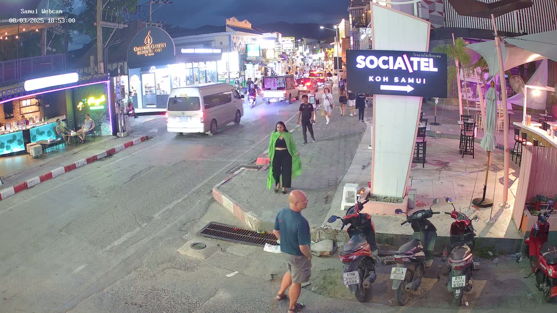 A nighttime street scene shows pedestrians walking past shops and restaurants, with several motorbikes parked nearby and a few cars driving on the road.
