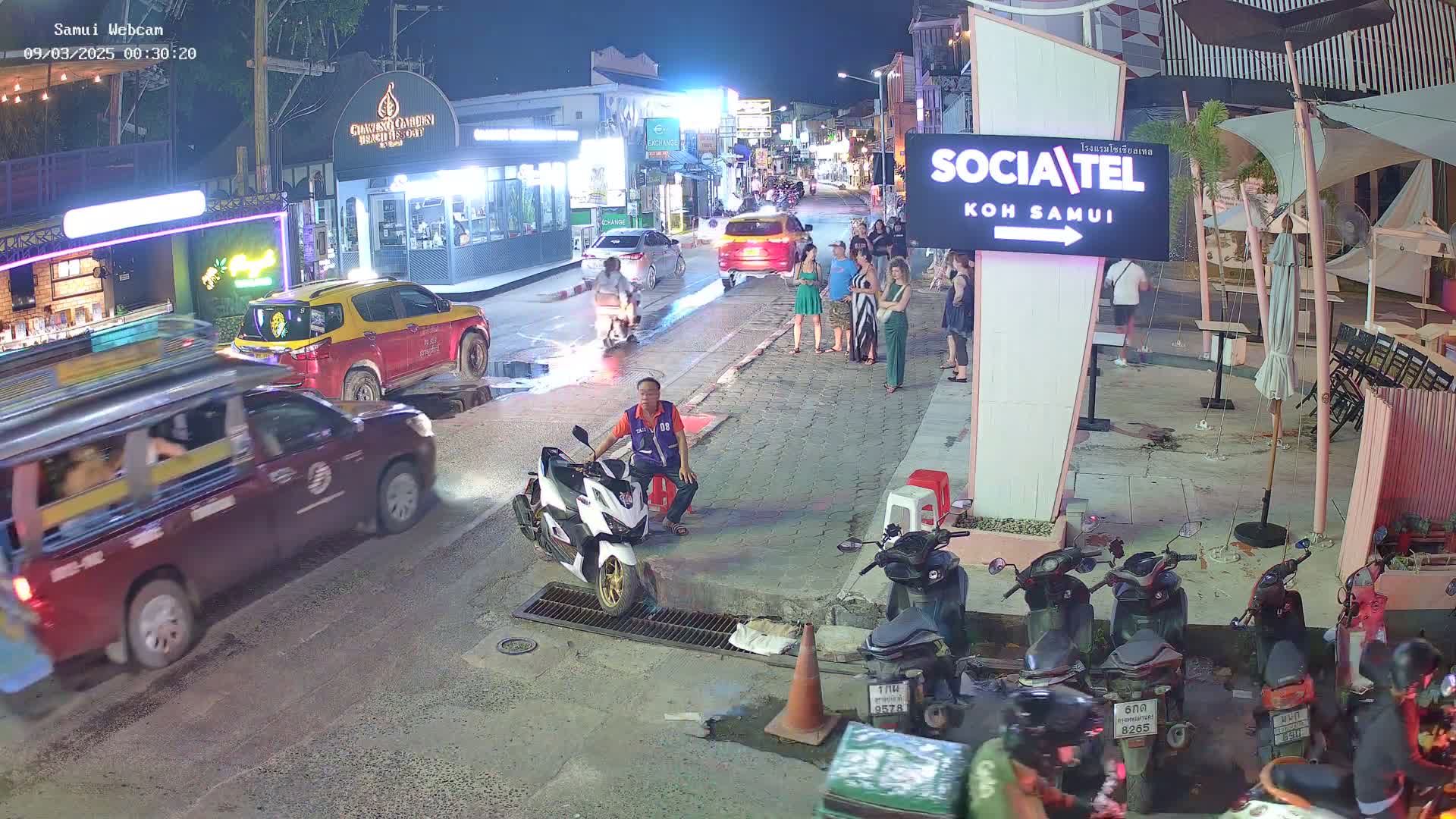 At night, a street scene shows several vehicles, including cars and motorcycles, parked and moving along a road lined with businesses and people standing outside.