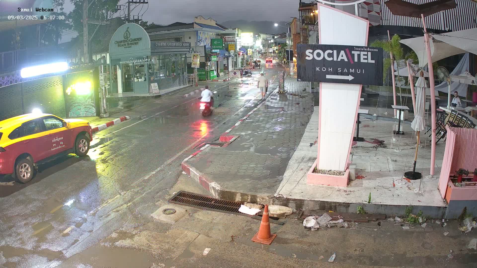 A wet street scene at night shows a taxi, a motorcyclist, and pedestrians near shops and a restaurant with outdoor seating.