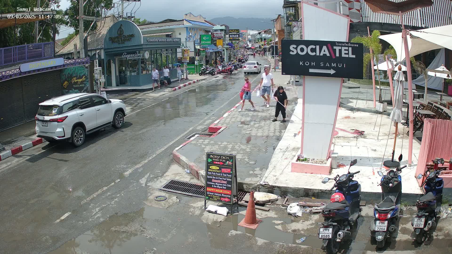A wet street scene shows pedestrians walking past shops and parked vehicles under a partly cloudy sky.