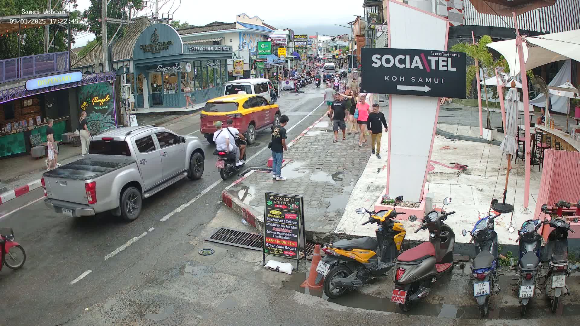 A wet street scene shows several vehicles, including a silver pickup truck and a yellow taxi, with pedestrians walking along a sidewalk past shops and restaurants under an overcast sky.