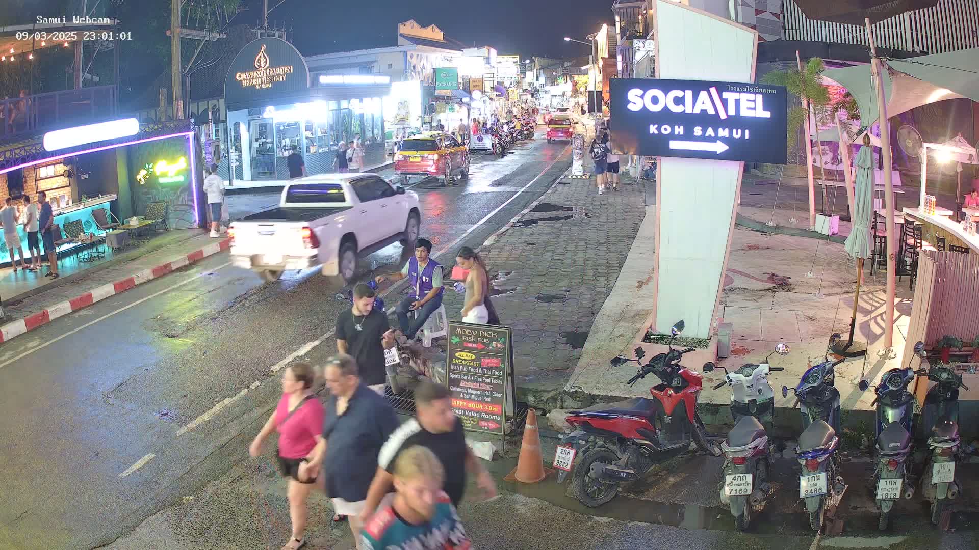 A wet, nighttime street scene shows pedestrians and vehicles, including several parked motorbikes and a white pickup truck, near various buildings and street vendors.