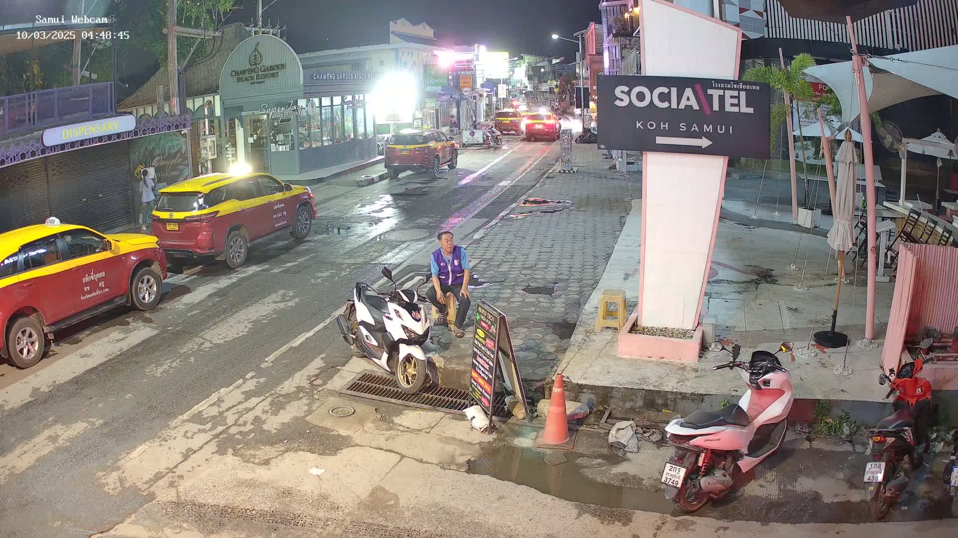 A man sits on a curb next to a parked scooter at night on a wet street lined with parked cars and buildings, under dim lighting.