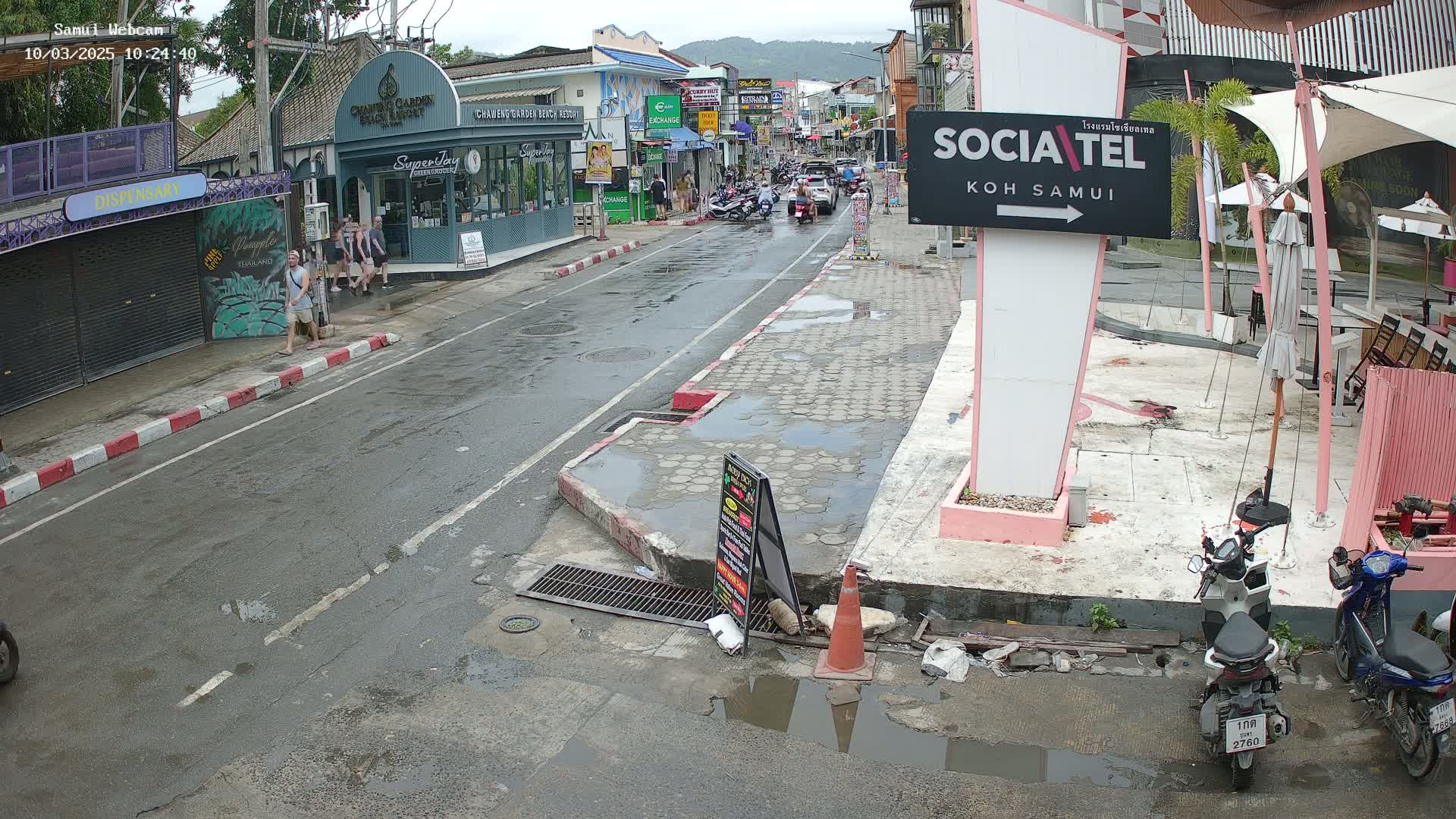 A wet street scene shows several people walking past shops and restaurants, with motorbikes parked along the side of the road, under an overcast sky.