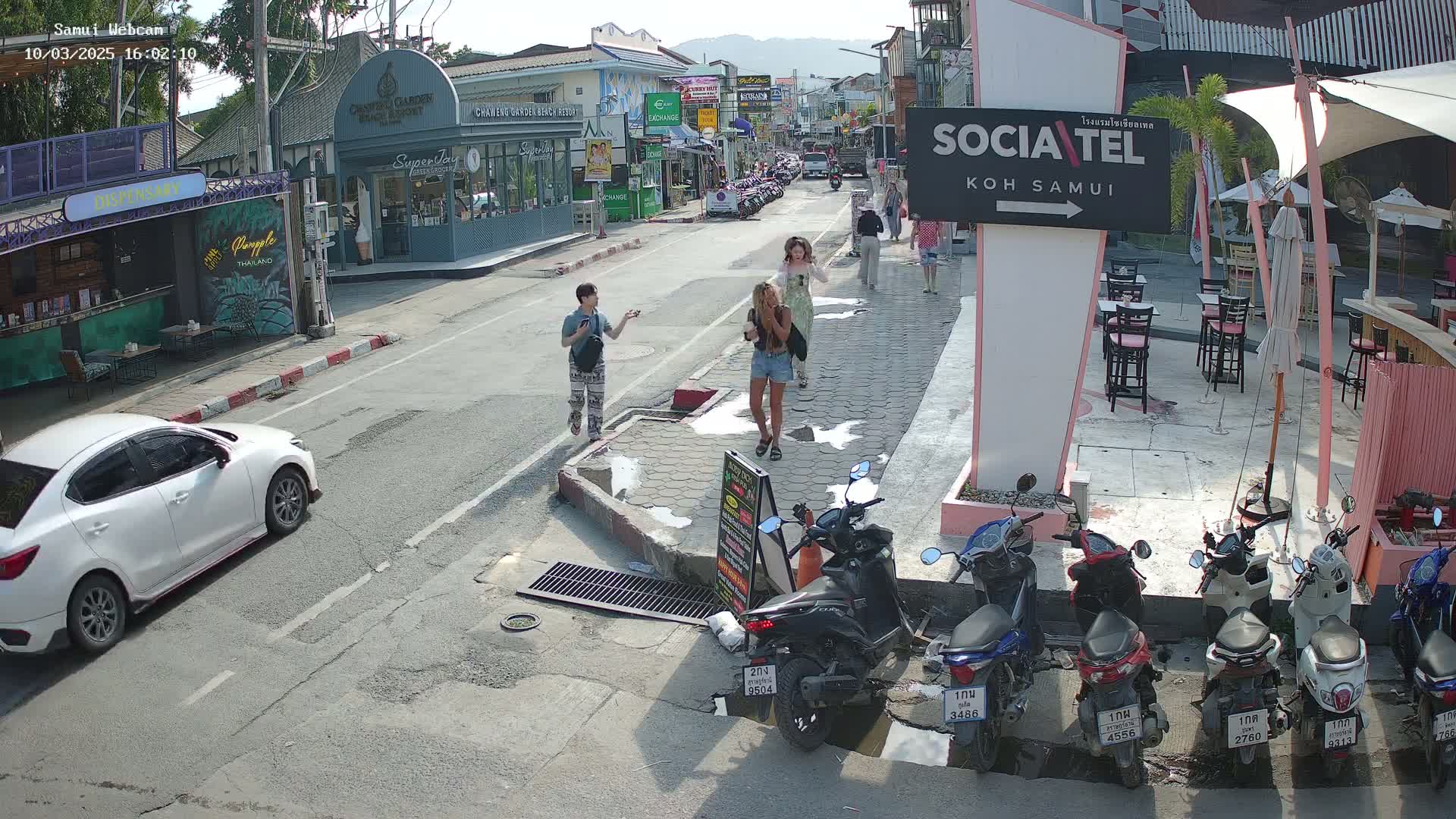 A sunny street scene shows several people walking past shops and restaurants, with several parked motor scooters and a white car in the foreground.