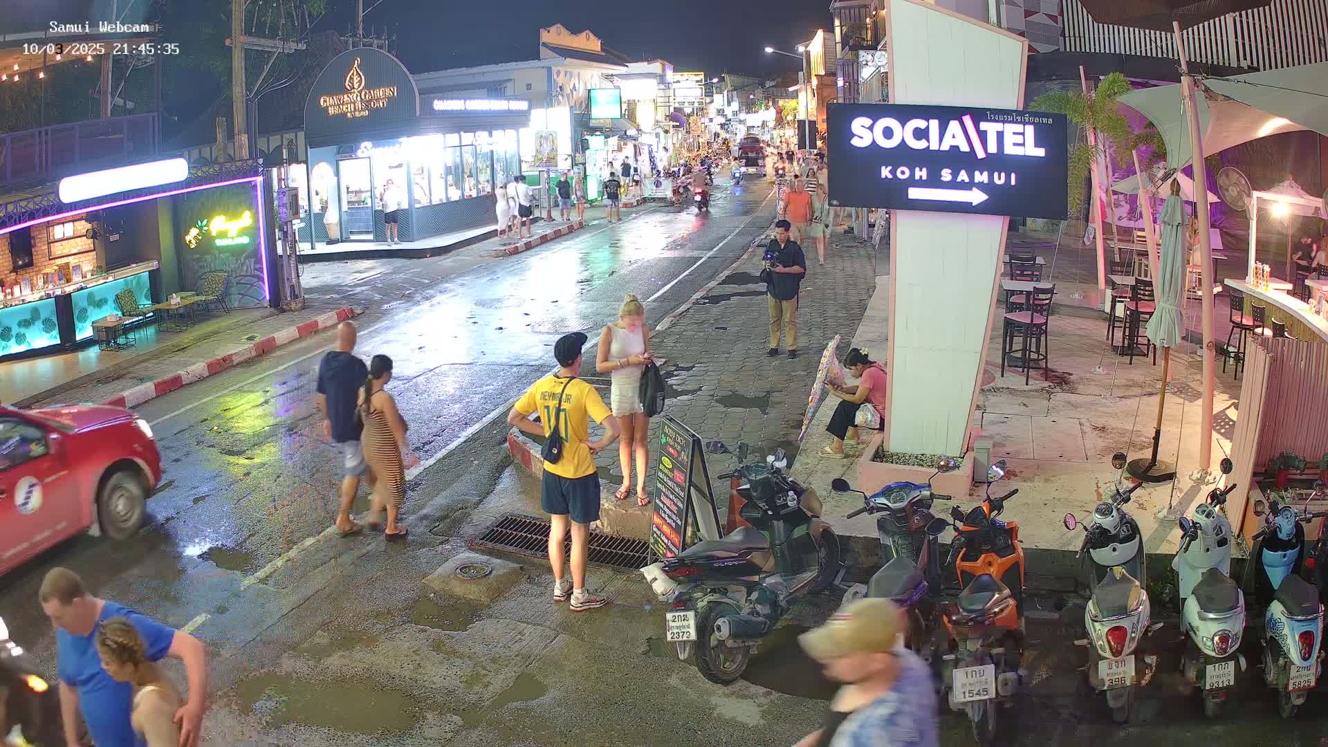 A wet street at night, lined with shops and restaurants, with people walking and motorbikes parked along the side.