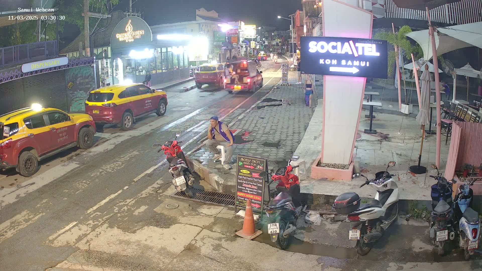 A nighttime street scene shows several parked taxis and motorbikes, a man sitting on a curb, and outdoor seating areas of establishments.