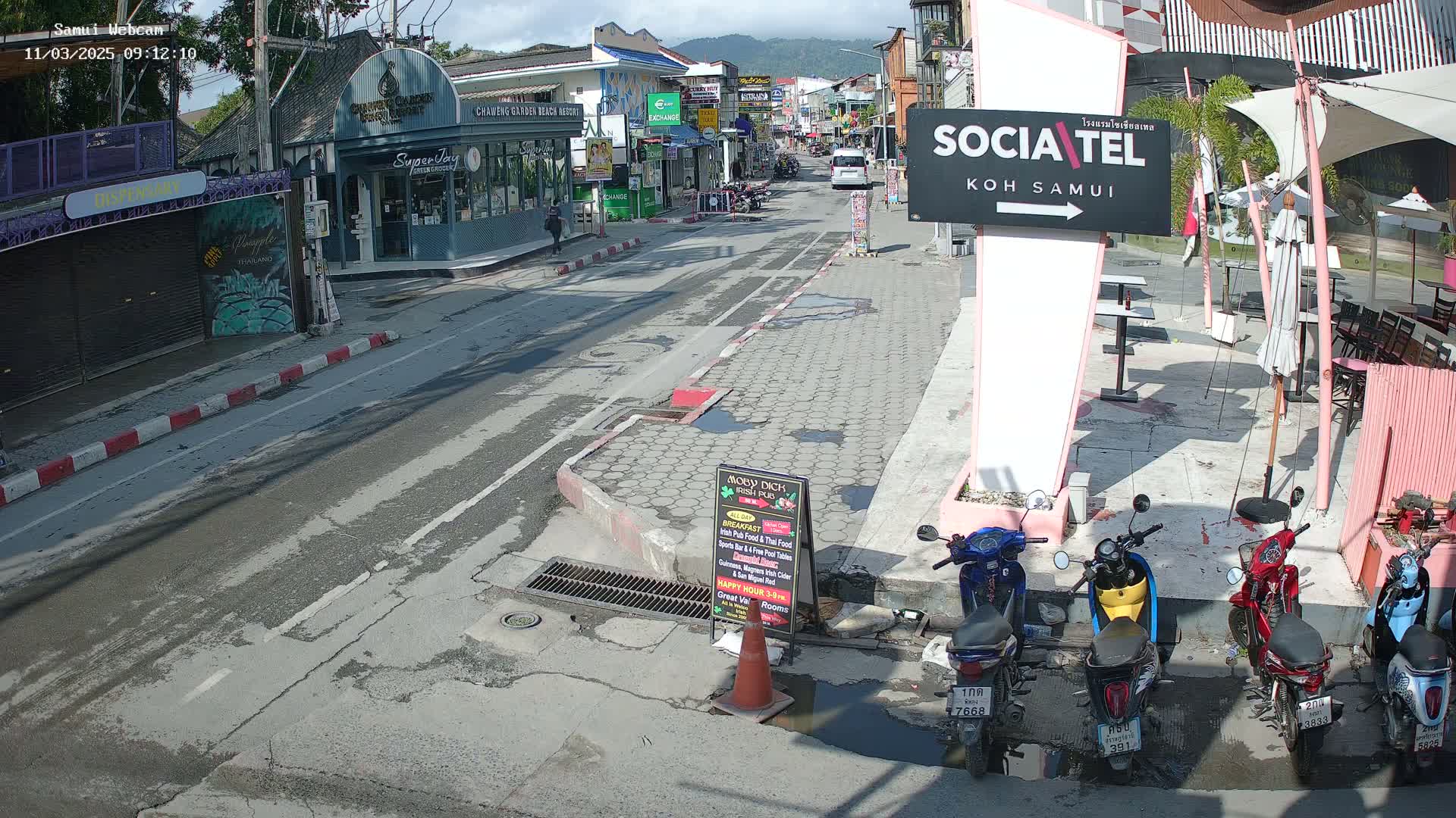 A sunny street scene shows a mostly empty road lined with shops and parked motorbikes, with mountains visible in the distance.