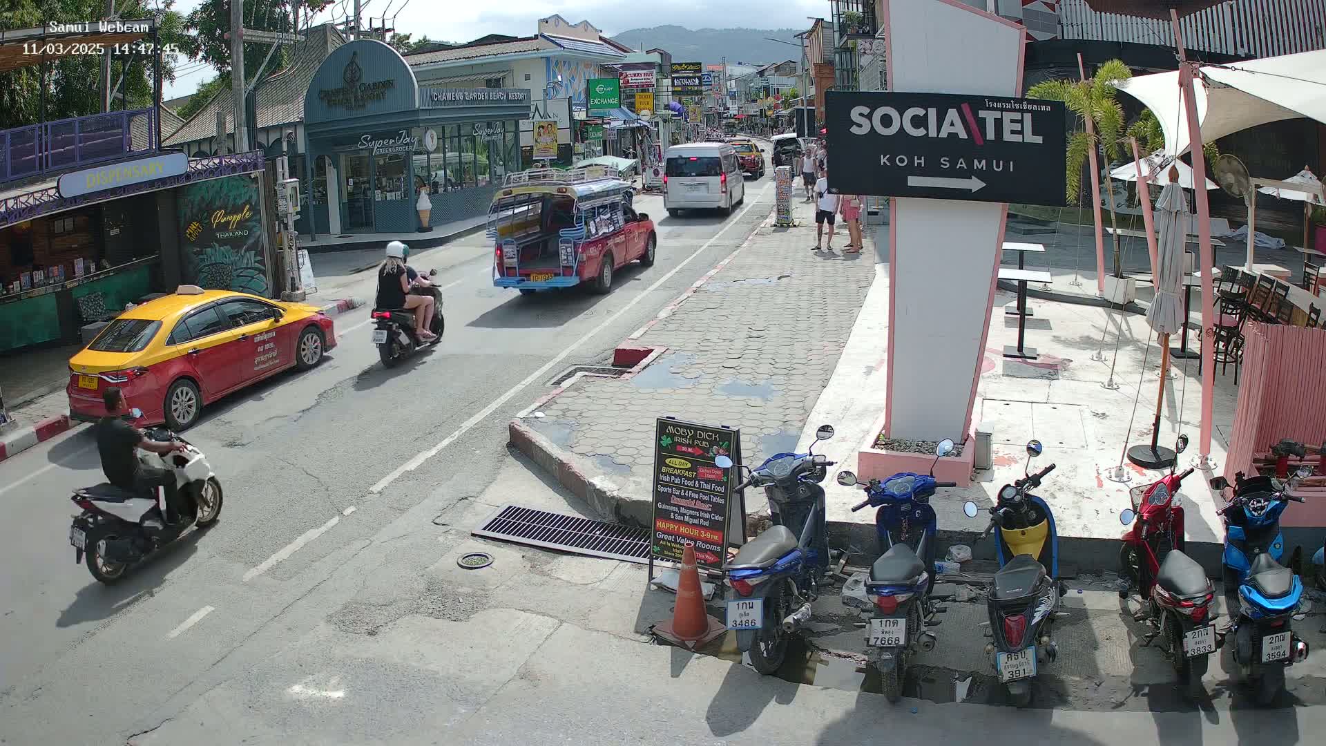 A sunny street scene in Koh Samui shows several motorbikes parked near outdoor seating, alongside a road with various vehicles including a taxi and songthaews, and pedestrians.