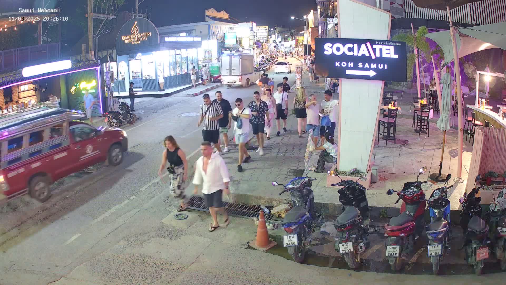 A nighttime street scene shows a group of pedestrians walking past parked motorbikes and a red pickup truck, under clear, dark skies.