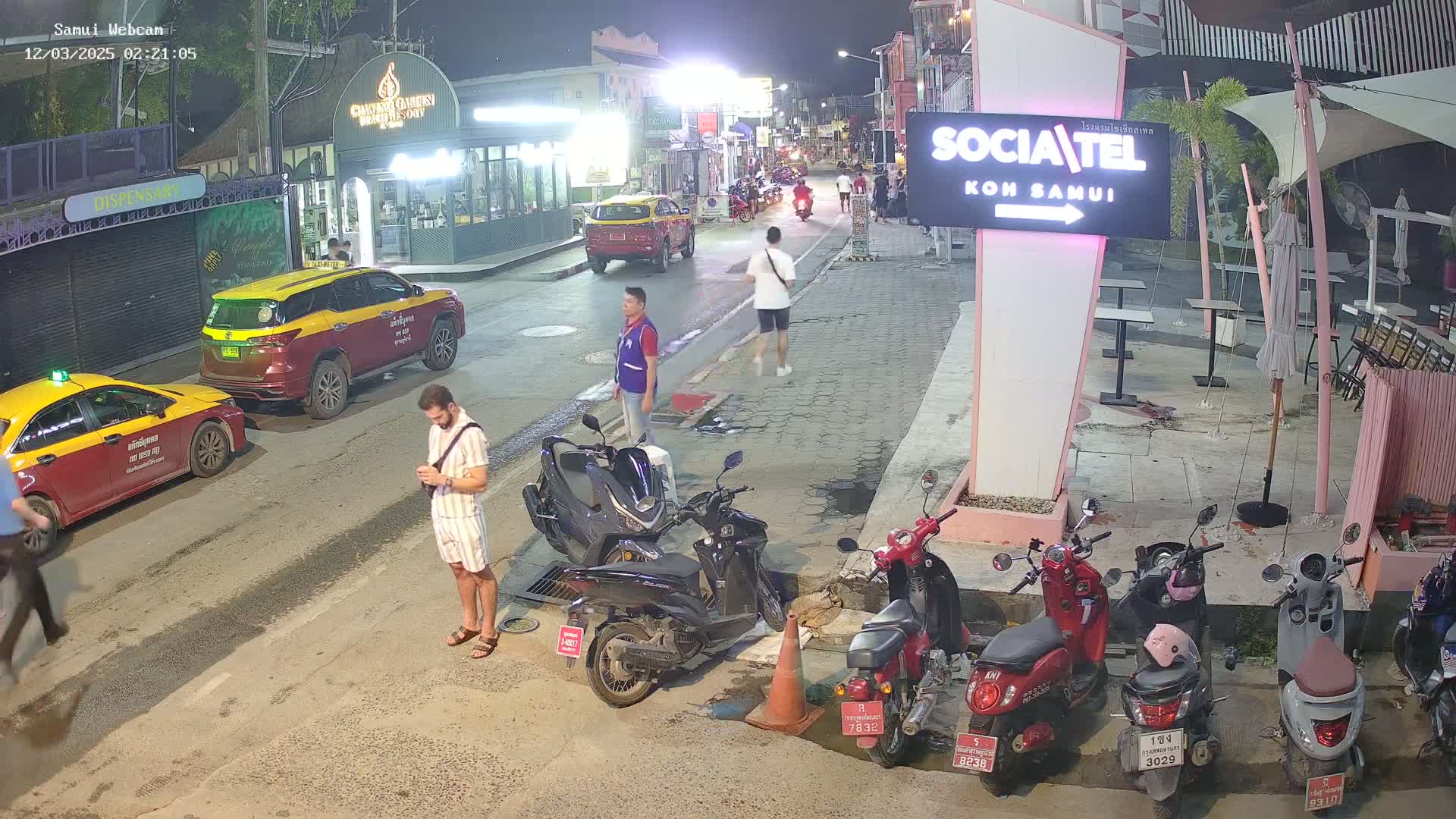 At night, a street scene shows several parked cars and motorbikes, a few pedestrians, and a man looking at his phone, all under clear night skies.