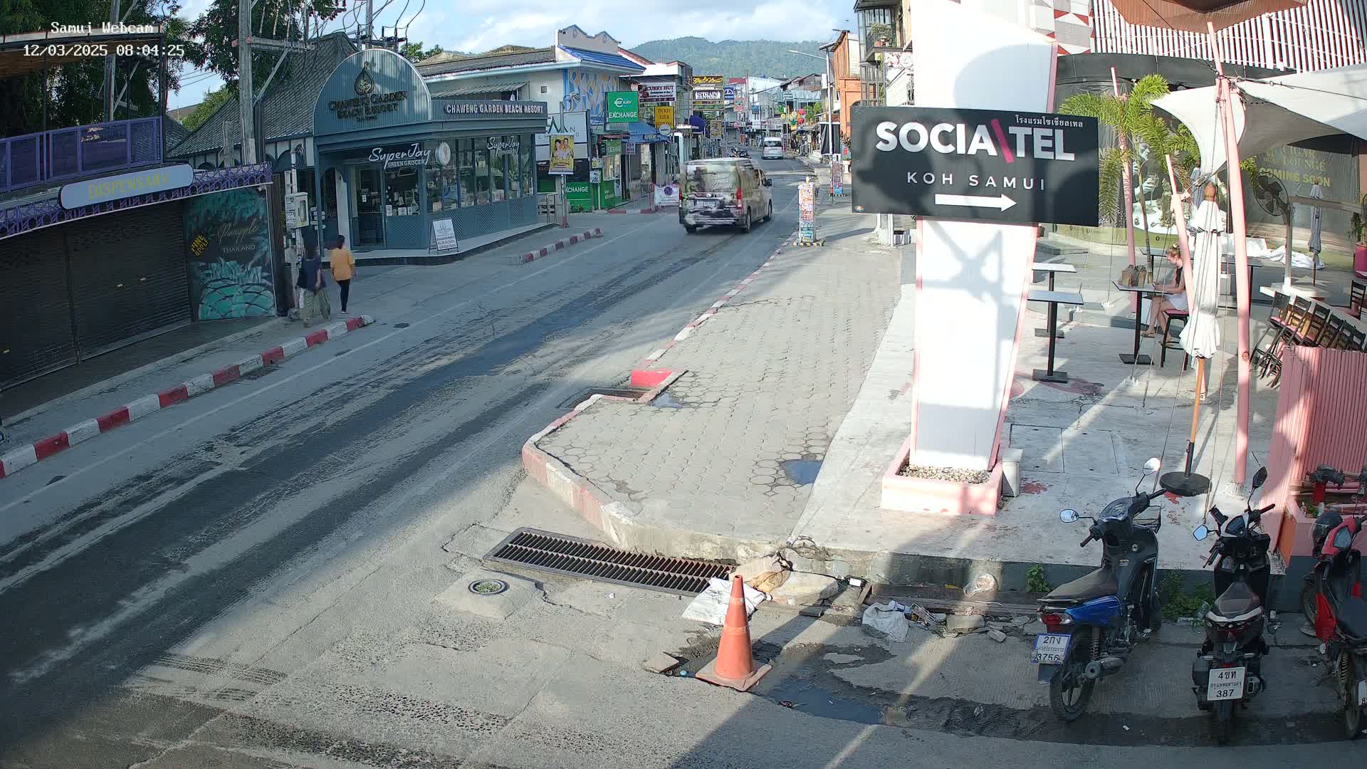 A sunny street scene shows a roadway with pedestrians, a van, and parked motorbikes, next to buildings with shops and a partially shaded outdoor seating area.