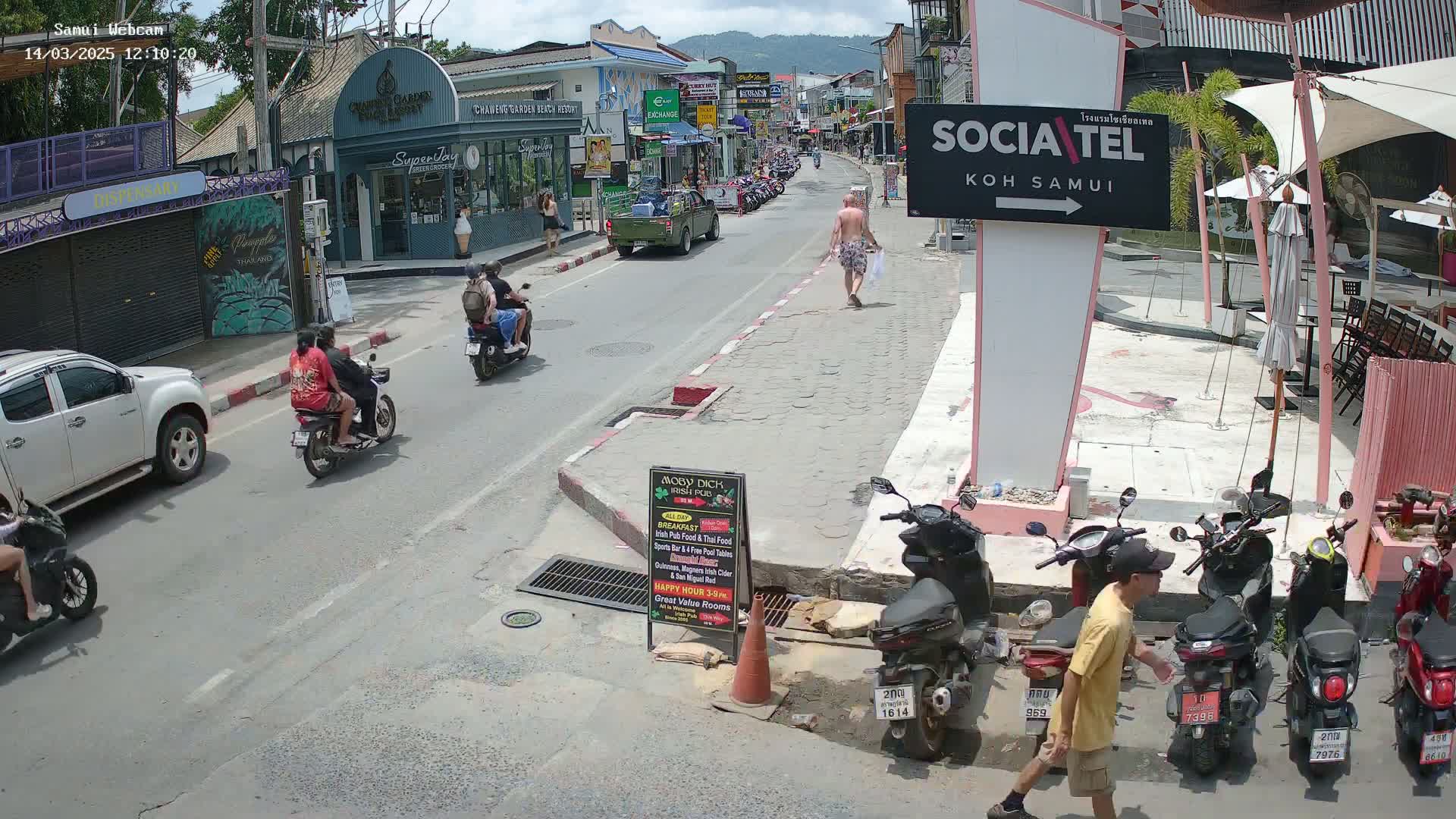 A sunny street scene shows several motorbikes and a few pedestrians, with cars parked along the roadside and buildings lining the street.