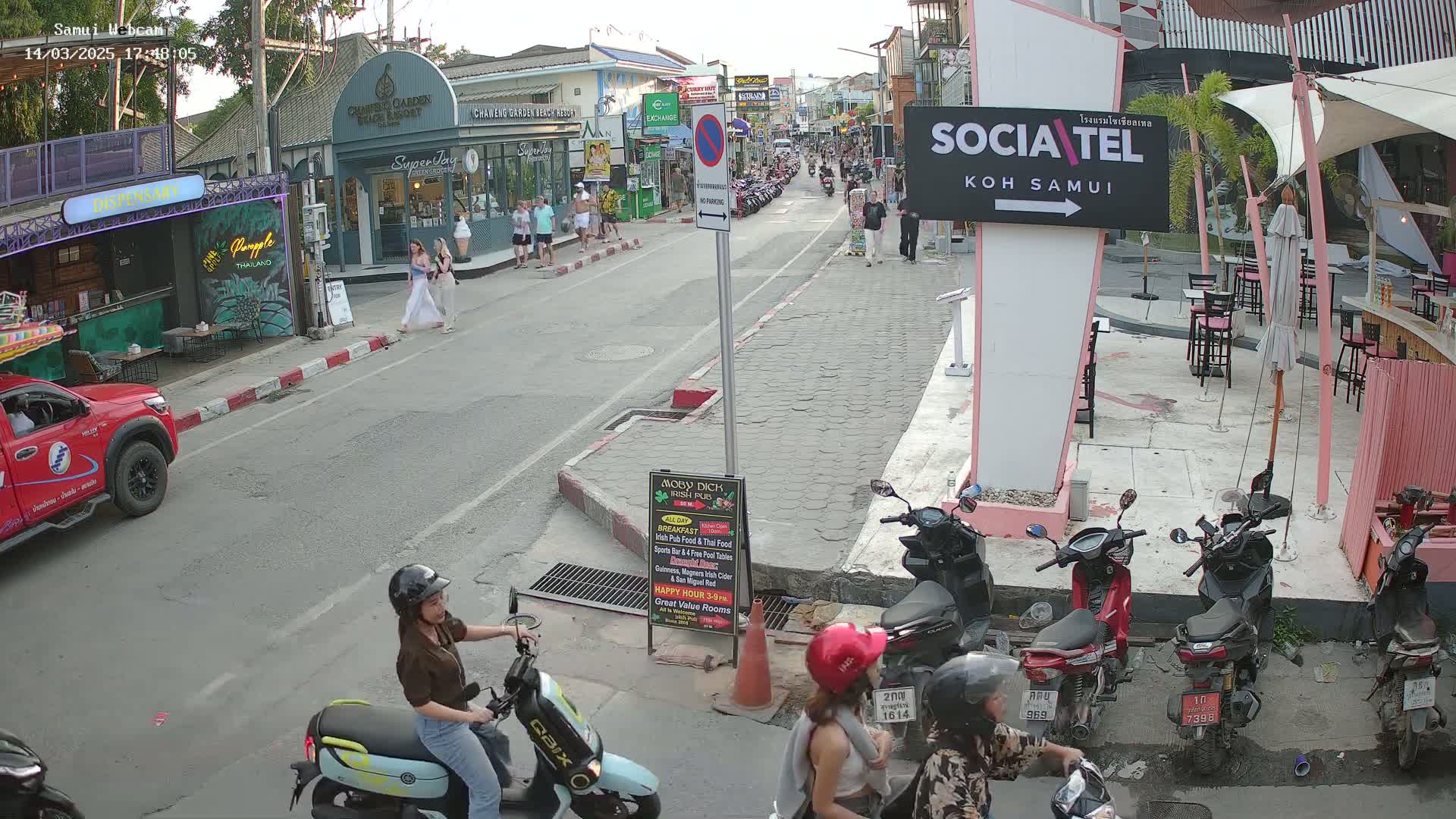 A street scene in what appears to be a Southeast Asian town shows several pedestrians, parked motorbikes, and a few people on scooters, under a partly cloudy sky.