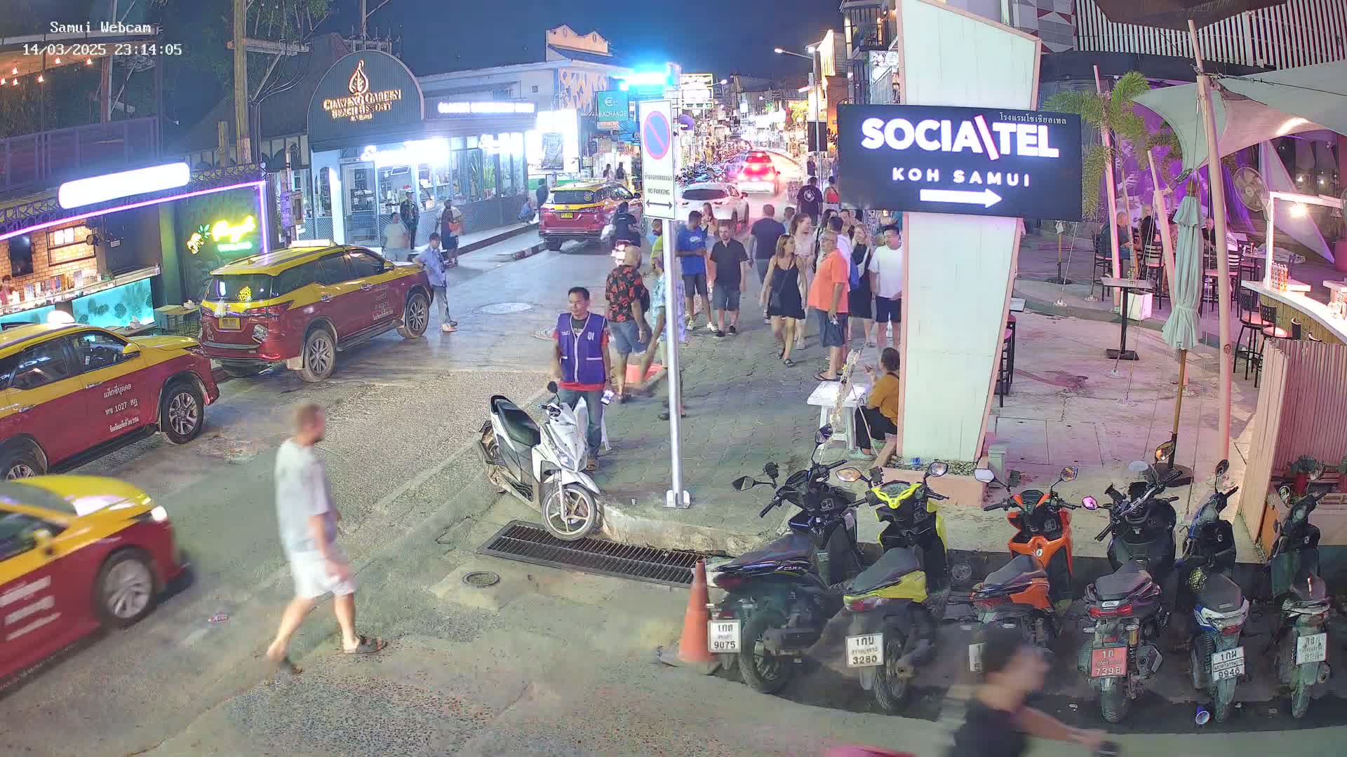 A nighttime street scene shows people walking and standing around parked cars and motorcycles, with several businesses visible in the background.
