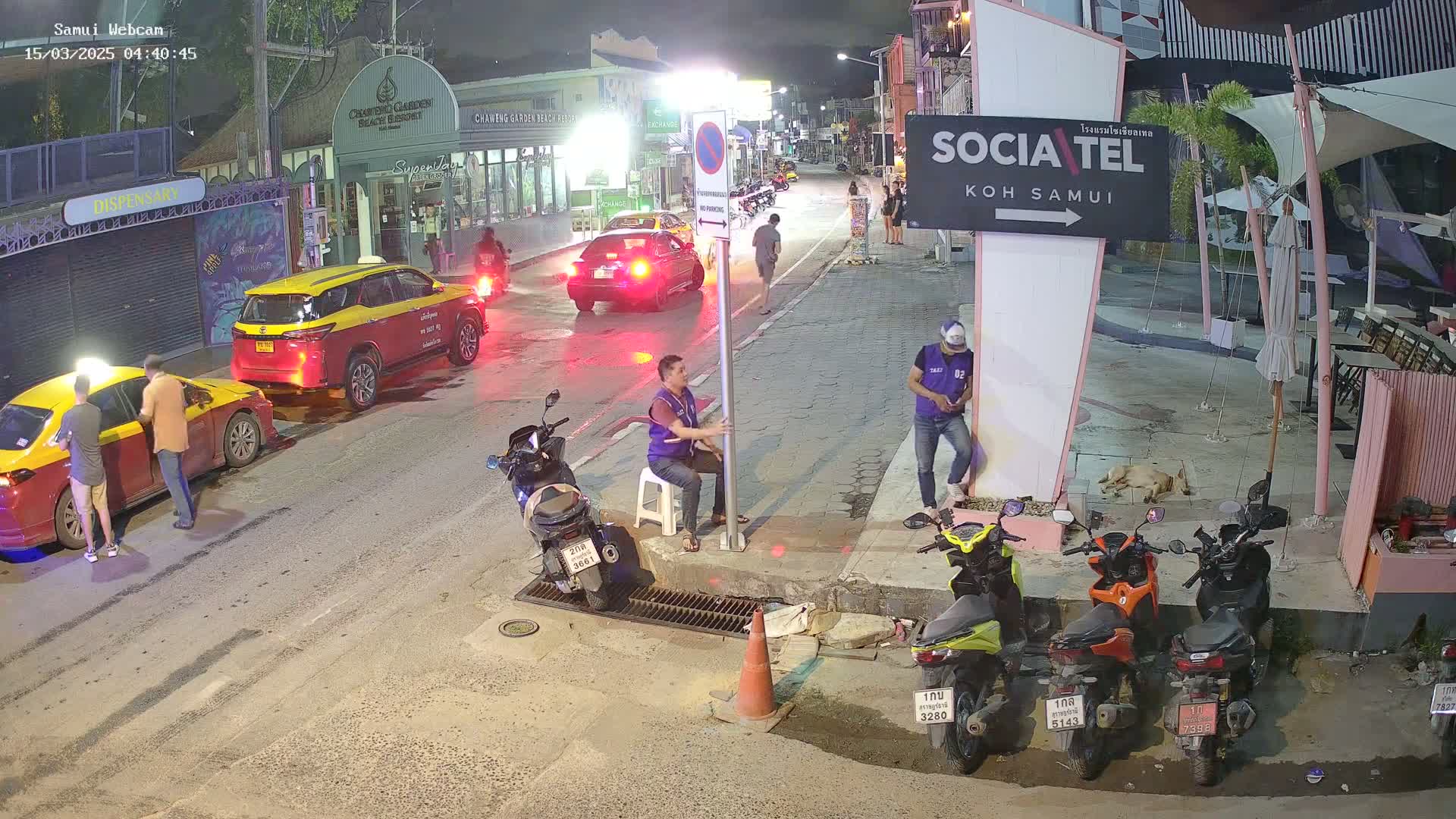 At night, a street scene shows several people standing near taxis and motorbikes parked along a curb, under outdoor lighting.