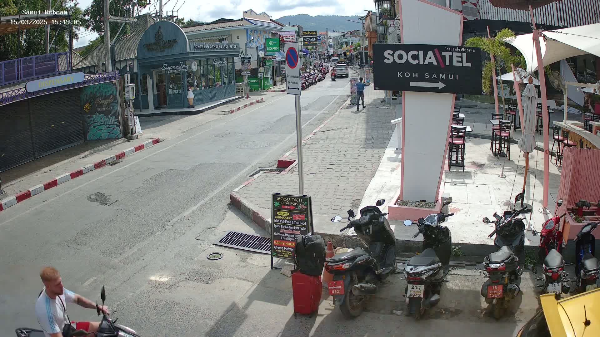 A sunny street scene shows a man on a scooter, parked motorcycles, outdoor seating at a cafe, and buildings lining a street.