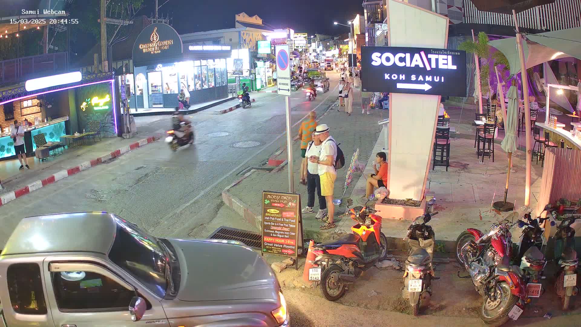 A nighttime street scene shows people walking and standing around parked motorcycles and cars near brightly lit shops and restaurants under a clear sky.