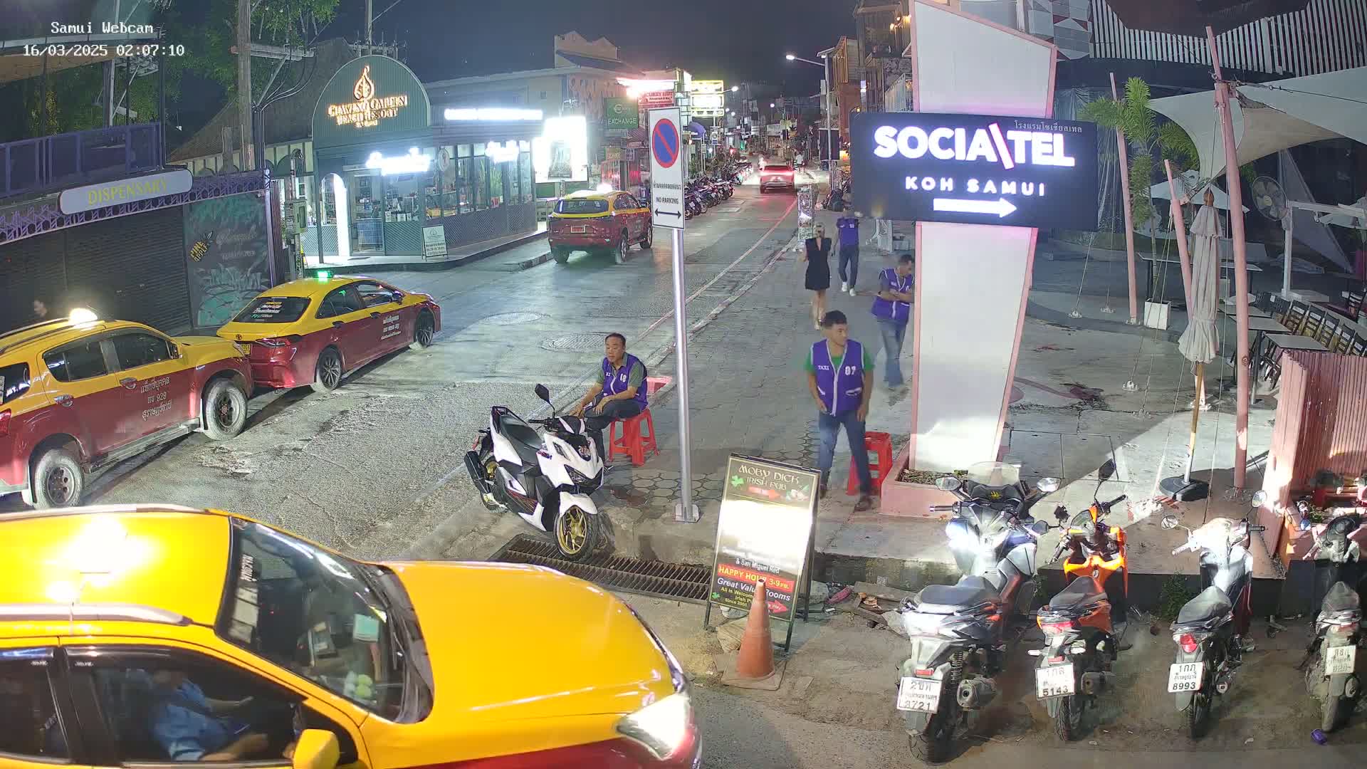 A nighttime street scene shows several yellow taxis, motor scooters, and people, with some people sitting and others standing near a building.