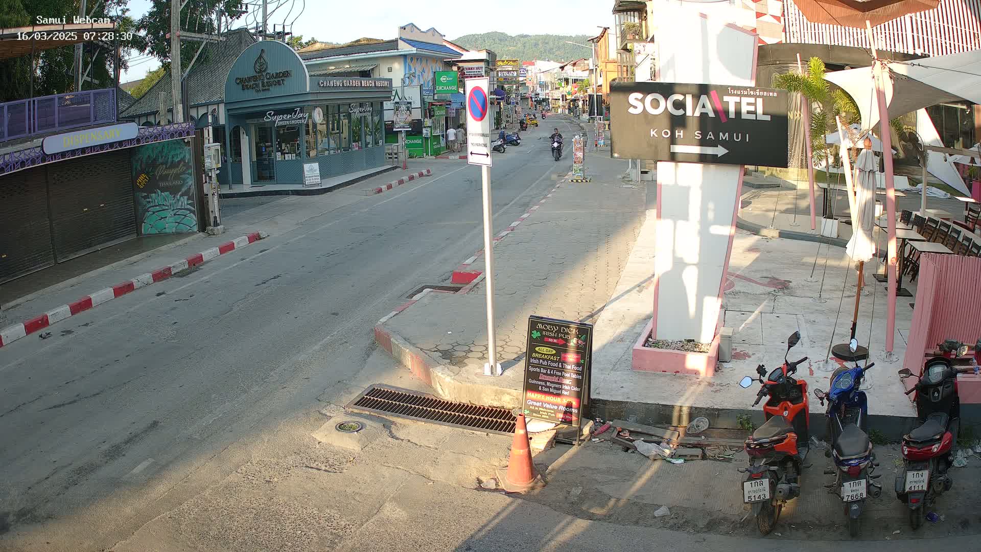 A mostly empty street scene in bright sunlight shows several parked motorbikes near a pink building, with shops and buildings lining the street, and a few people walking or riding motorbikes in the distance.