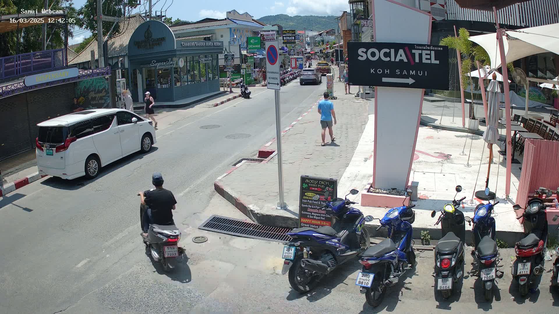 A sunny street scene shows a white minivan parked on the side of the road, several people walking, a man on a scooter, and numerous parked motorbikes in front of various buildings.