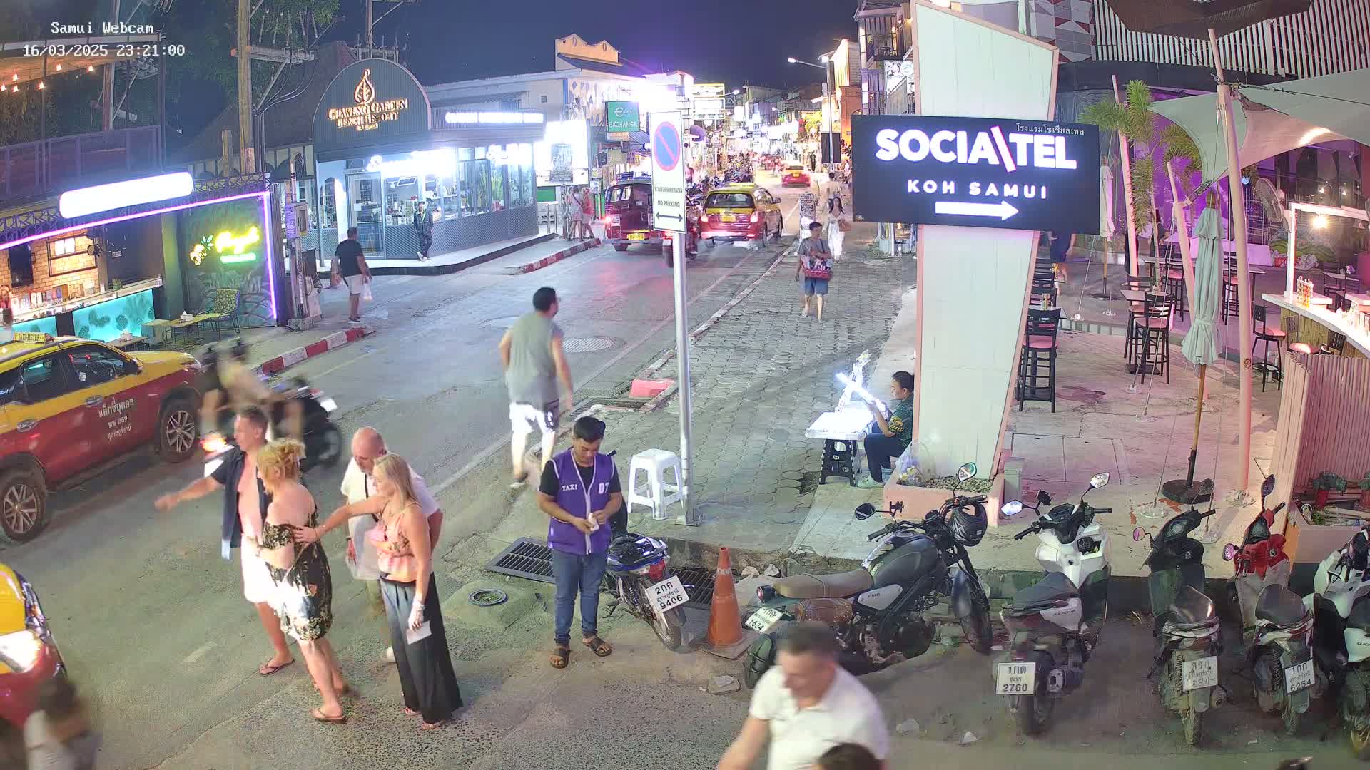 A nighttime street scene shows several people walking and standing near parked motorcycles and taxis, with brightly lit shops and restaurants in the background under a clear sky.